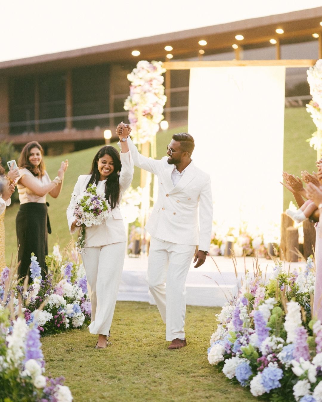 Smiling couple in matching white outfits walk hand in hand down a flower-lined aisle as guests cheer at an outdoor wedding ceremony.