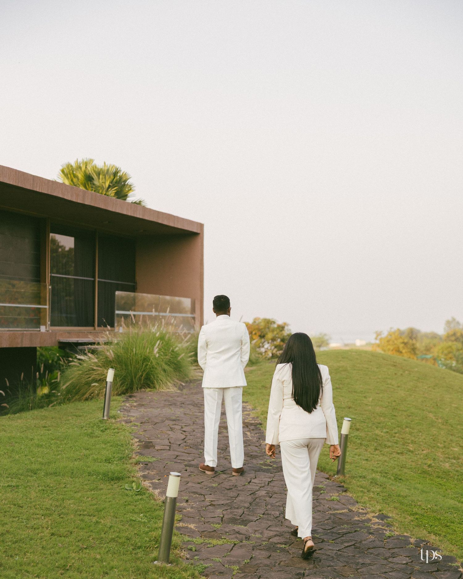 Couple in matching white outfits walk along a stone path through green grass toward a modern wooden-and-glass building at sunset.