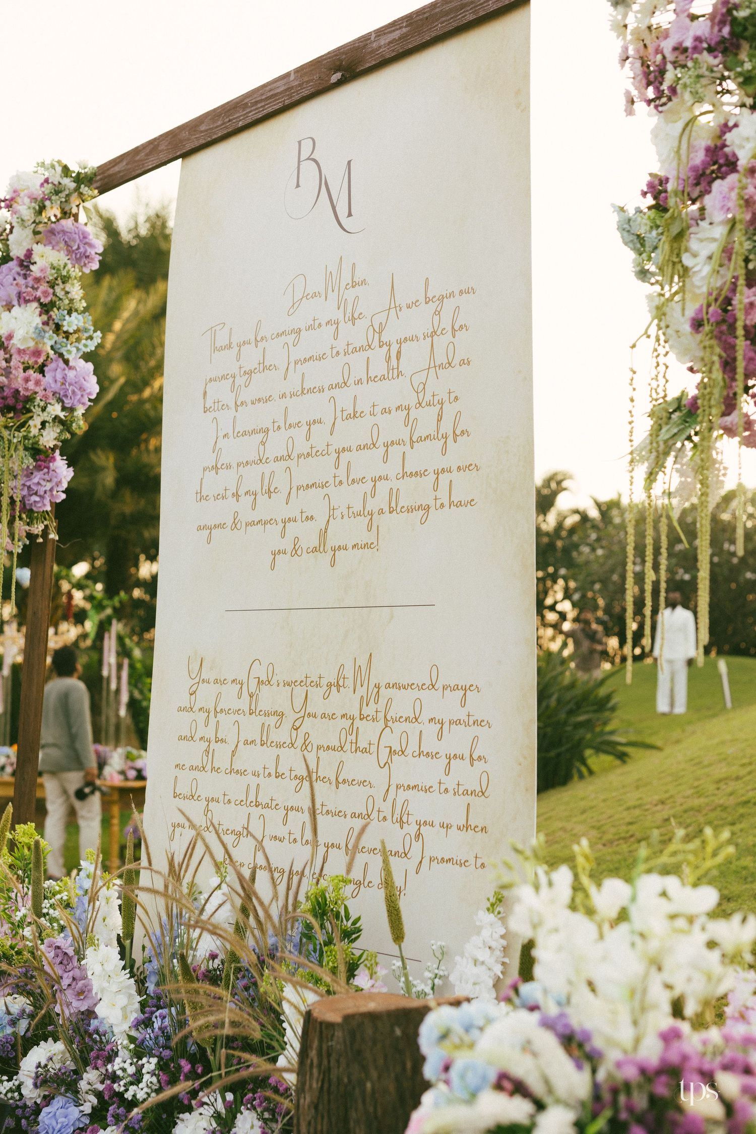 Elegant outdoor wedding ceremony sign with gold calligraphy, framed by lush pastel purple and white floral arrangements on a sunny lawn.