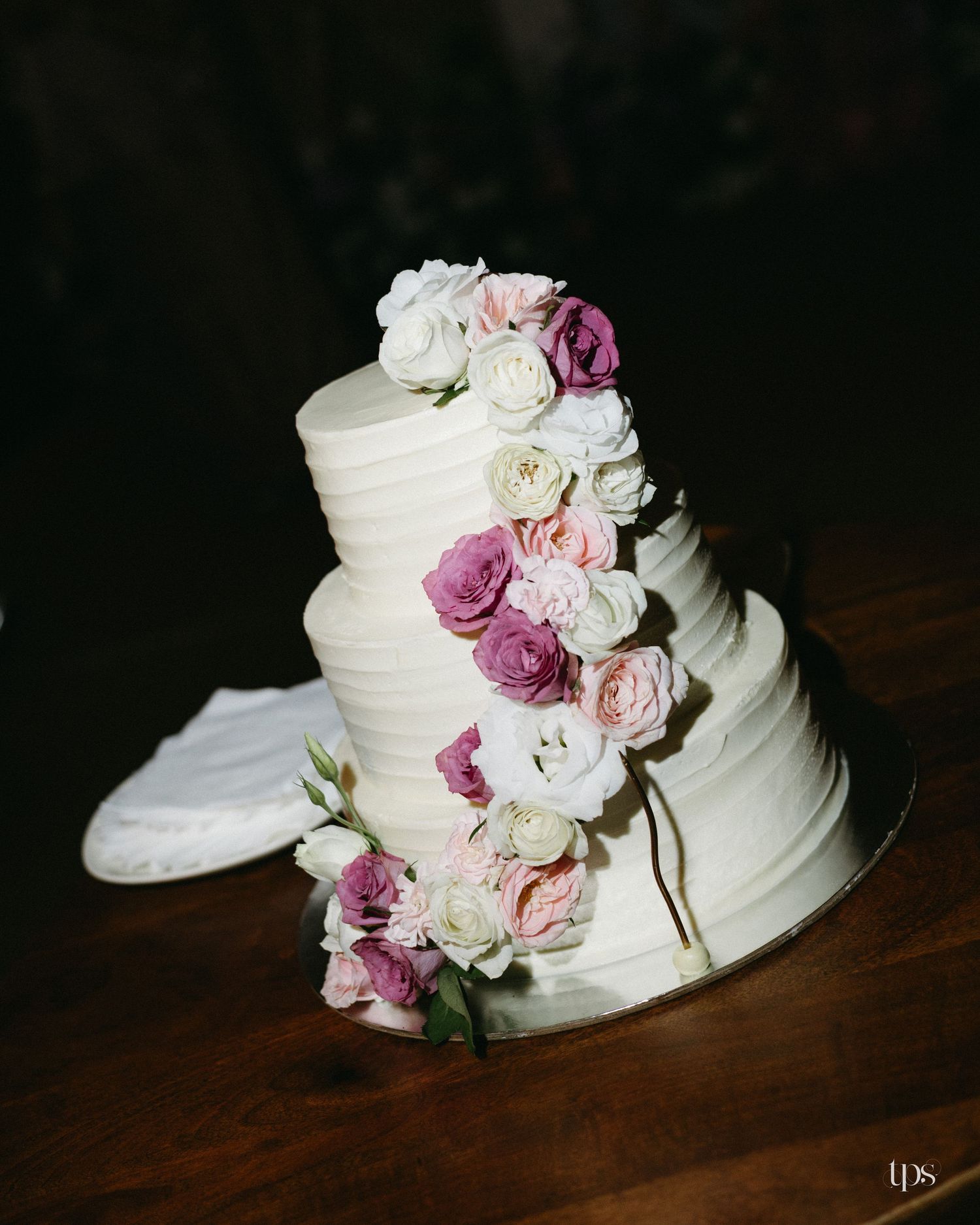 Three-tier white buttercream wedding cake with a cascade of pink, mauve and ivory roses on a mirrored board atop a rustic wooden table.