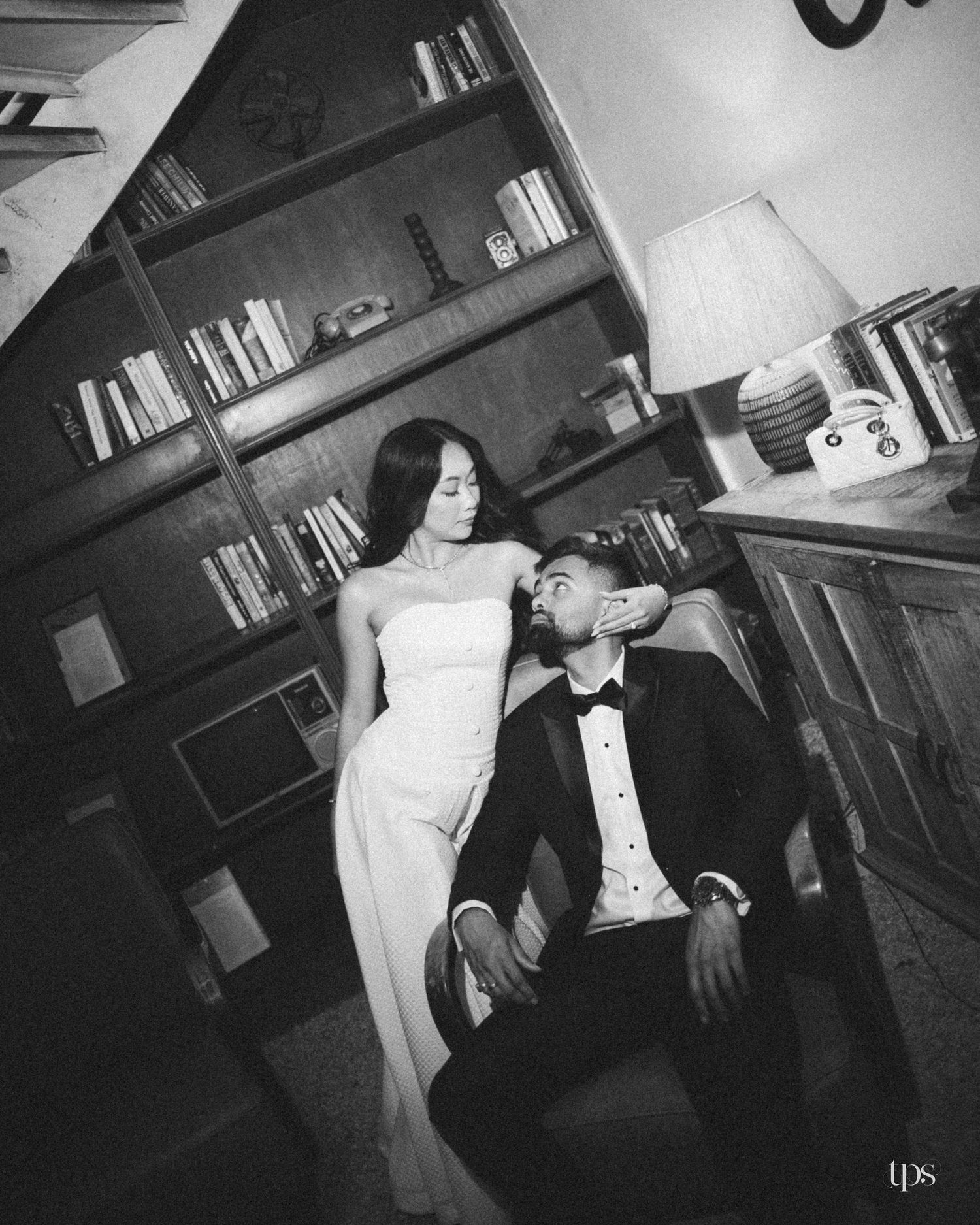 Black and white photo of an elegant couple in formal wear posing together beside a vintage bookshelf and lamp in a cozy study.