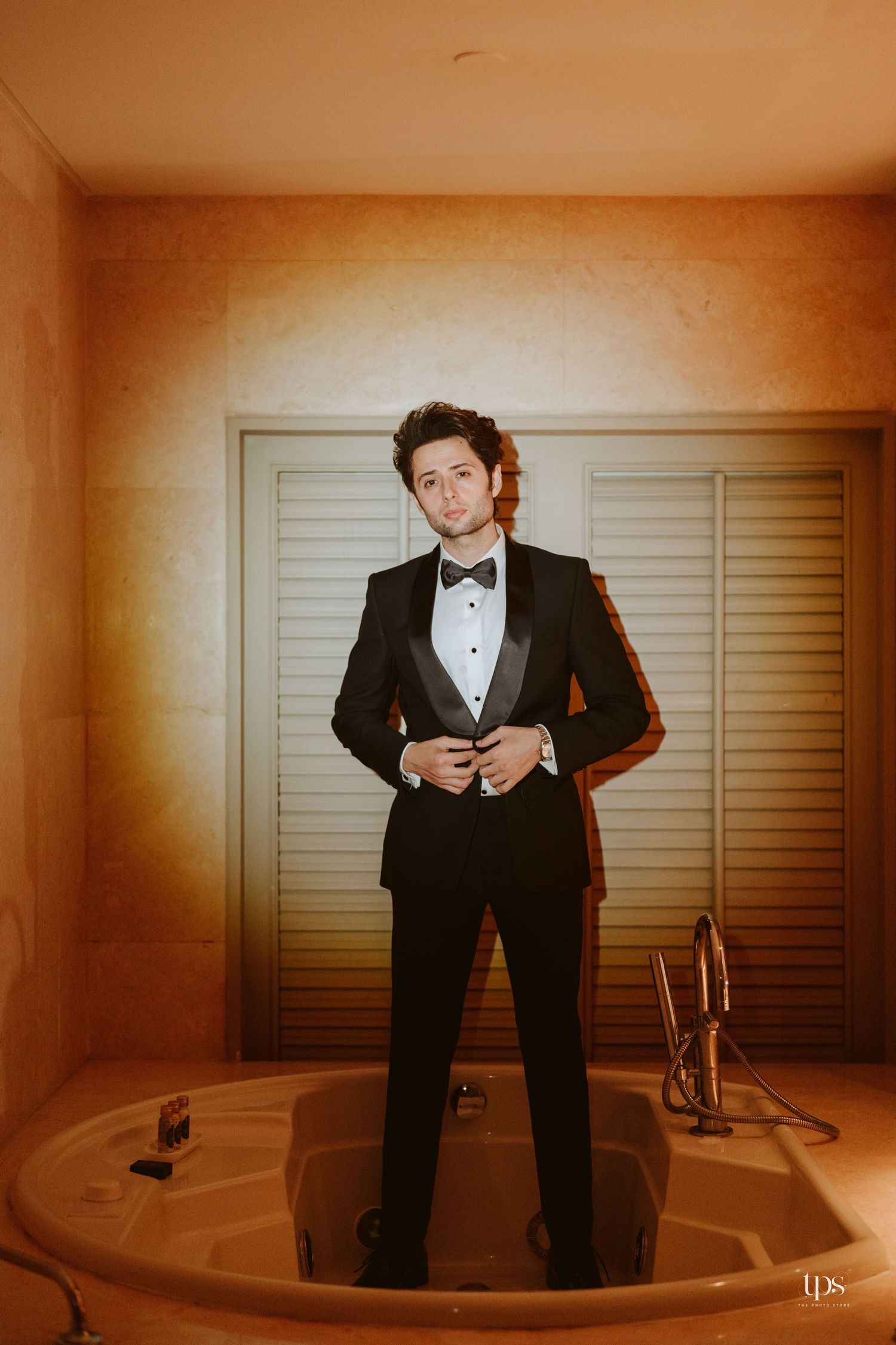 Groom in black tuxedo with bow tie standing in elegant bathroom with warm lighting.