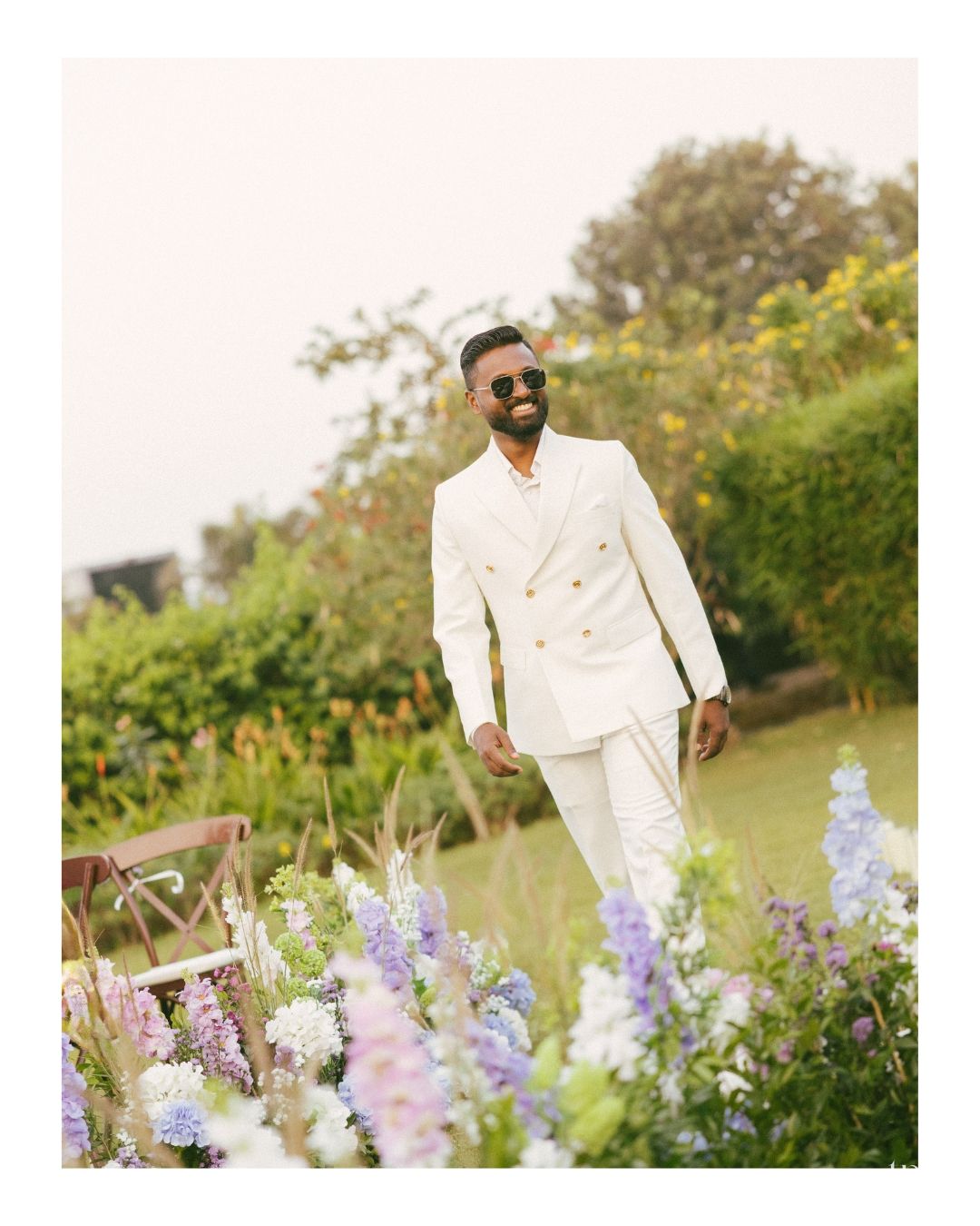 Man in a white double-breasted suit and sunglasses walks through a sunlit garden with blue and white delphiniums in the foreground.