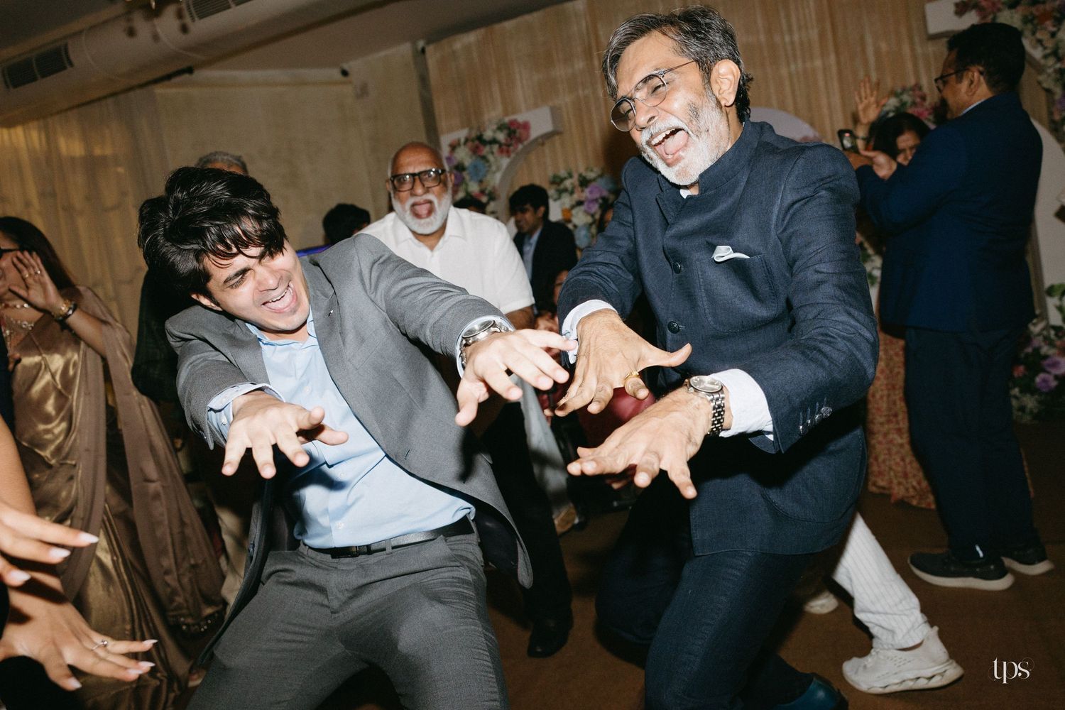 Two men in formal attire dance joyfully with outstretched arms at a lively indoor celebration while guests look on in the background.