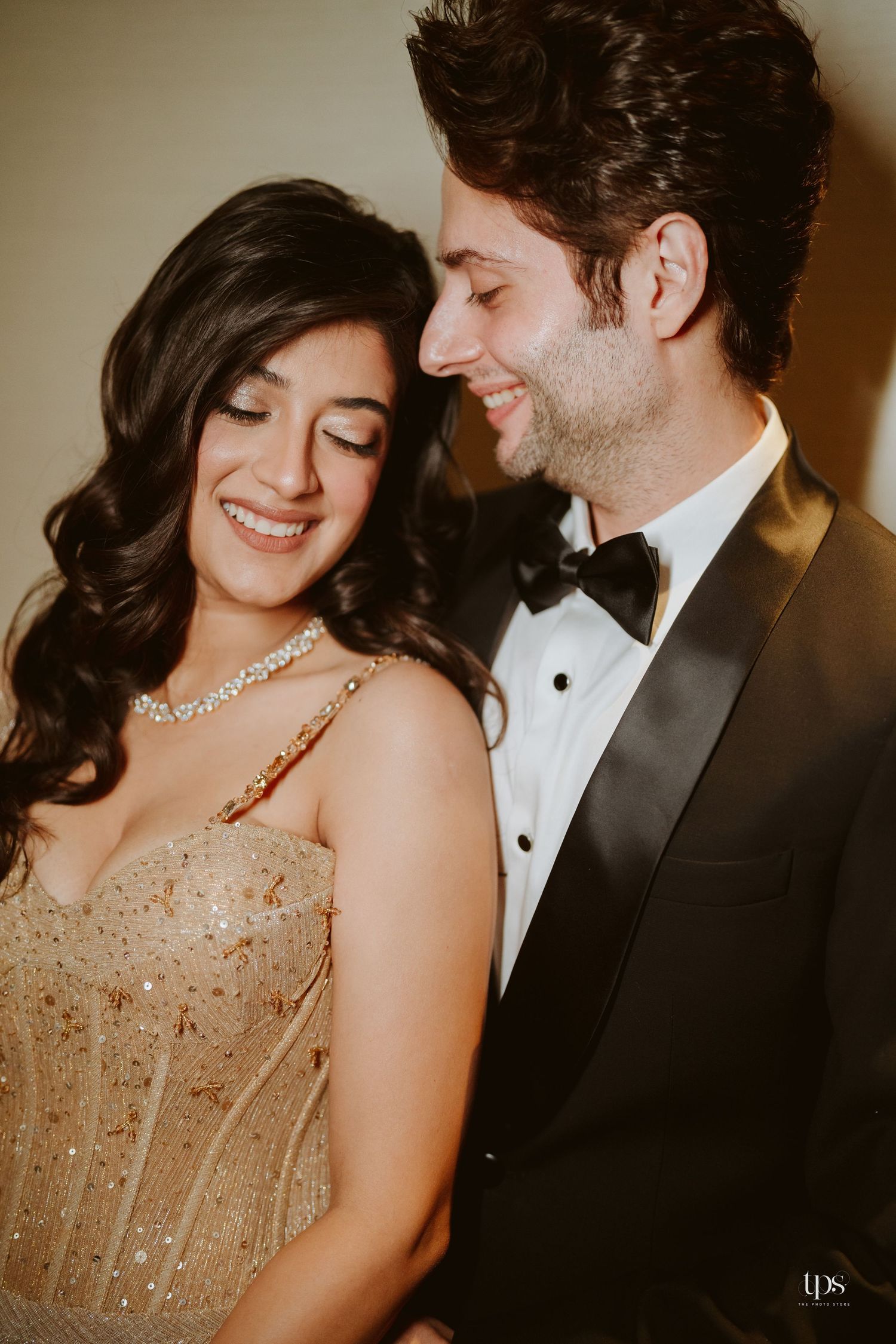 Elegant couple in formal attire, woman in gold beaded gown with diamond necklace, man in black tuxedo, sharing a joyful moment.