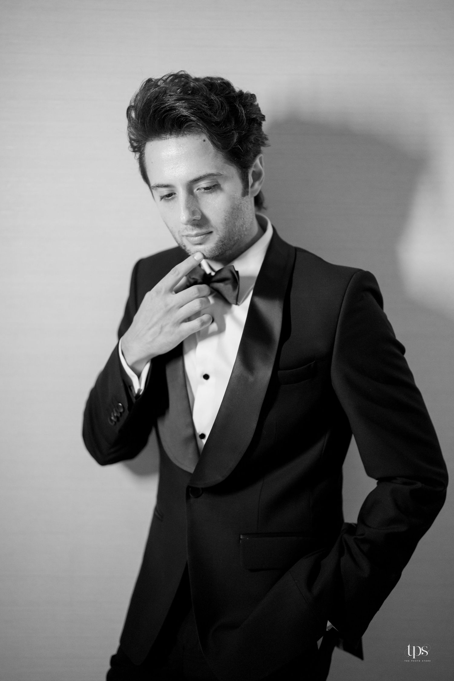 Black and white photo of a young man in a tuxedo adjusting his bow tie, posing elegantly against a light background.