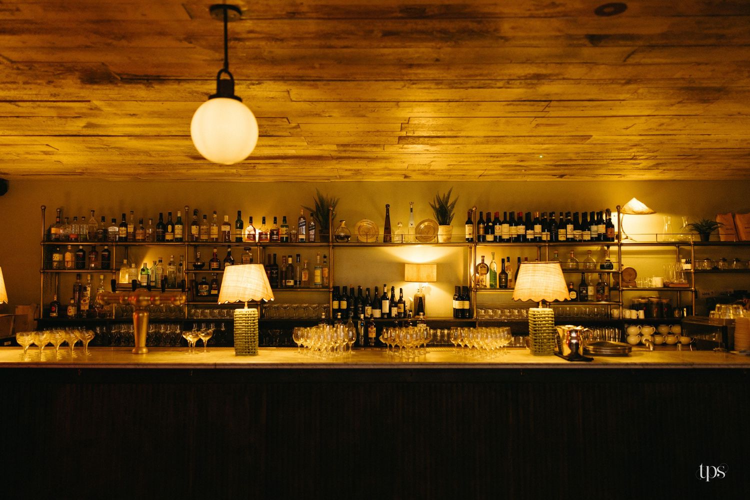 Warmly lit bar interior with shelves of bottles, glowing lamps, and a wooden ceiling in a cozy restaurant setting.