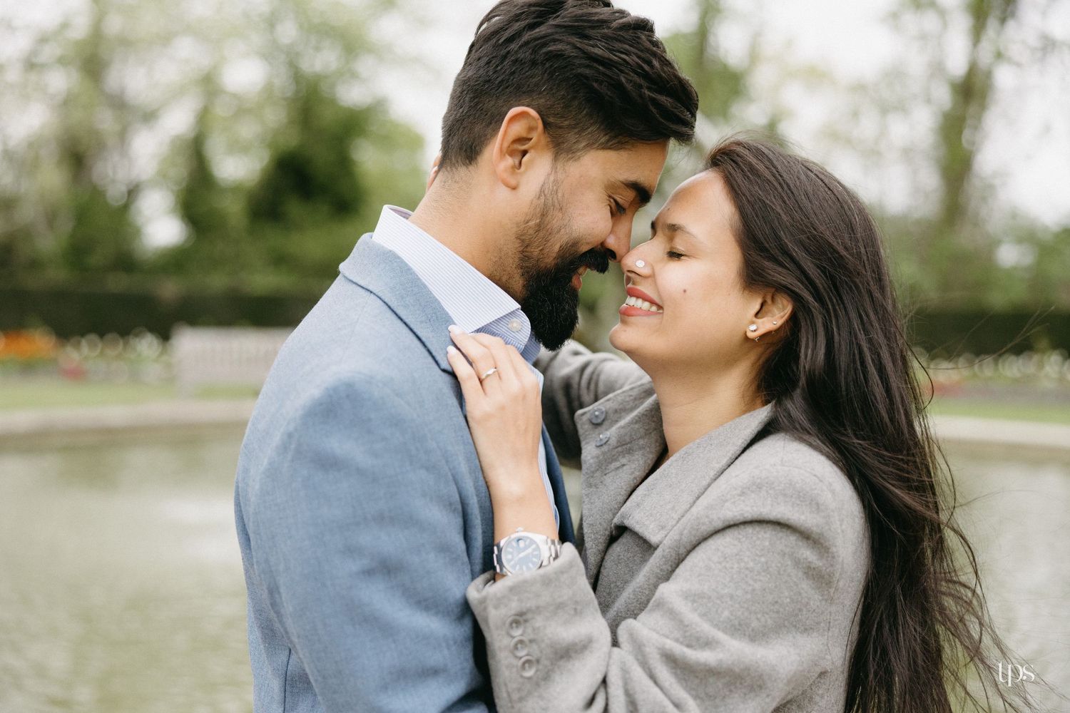 A couple shares a romantic moment near a garden fountain, the woman smiling as they lean close together outdoors.