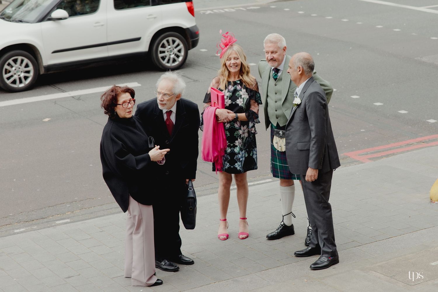 Five elegantly dressed people socializing on a city street, one man wearing a Scottish kilt at a formal outdoor gathering.