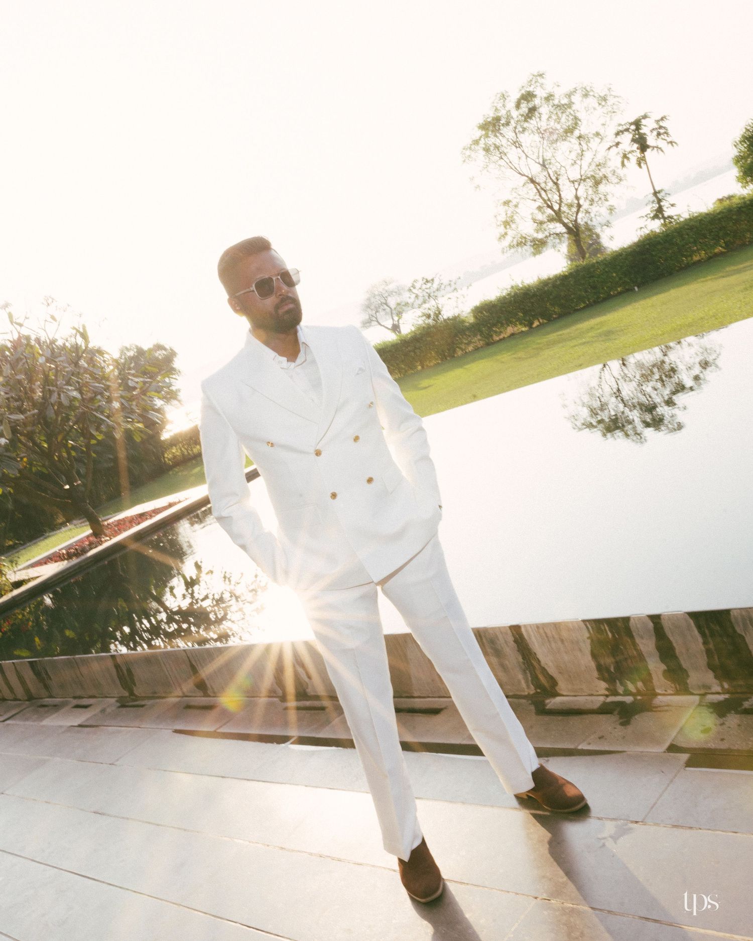 Man in a white double-breasted suit and sunglasses poses on a stone terrace beside a reflecting pool, with warm sunlight flaring behind him.
