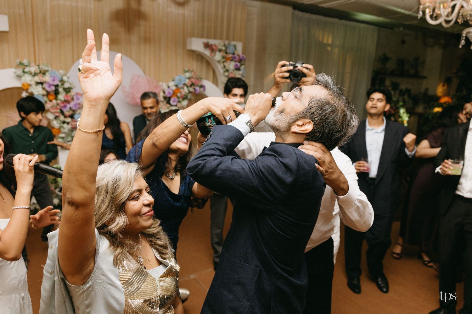 Wedding guests dance and celebrate at a reception as a man in a navy suit raises his arms while a photographer captures the lively moment.