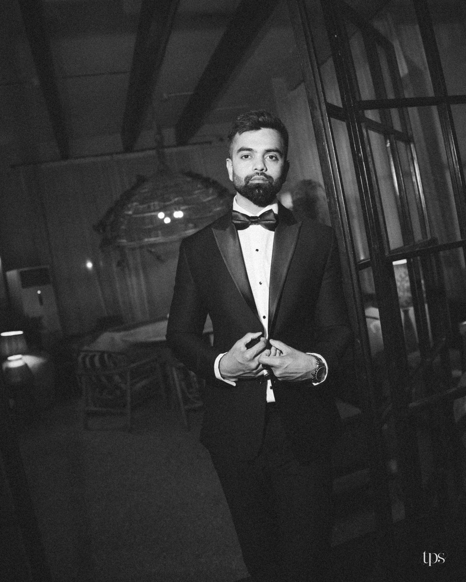 Black and white portrait of a bearded man in a tuxedo and bow tie adjusting his jacket in a dimly lit rustic room with glass doors.