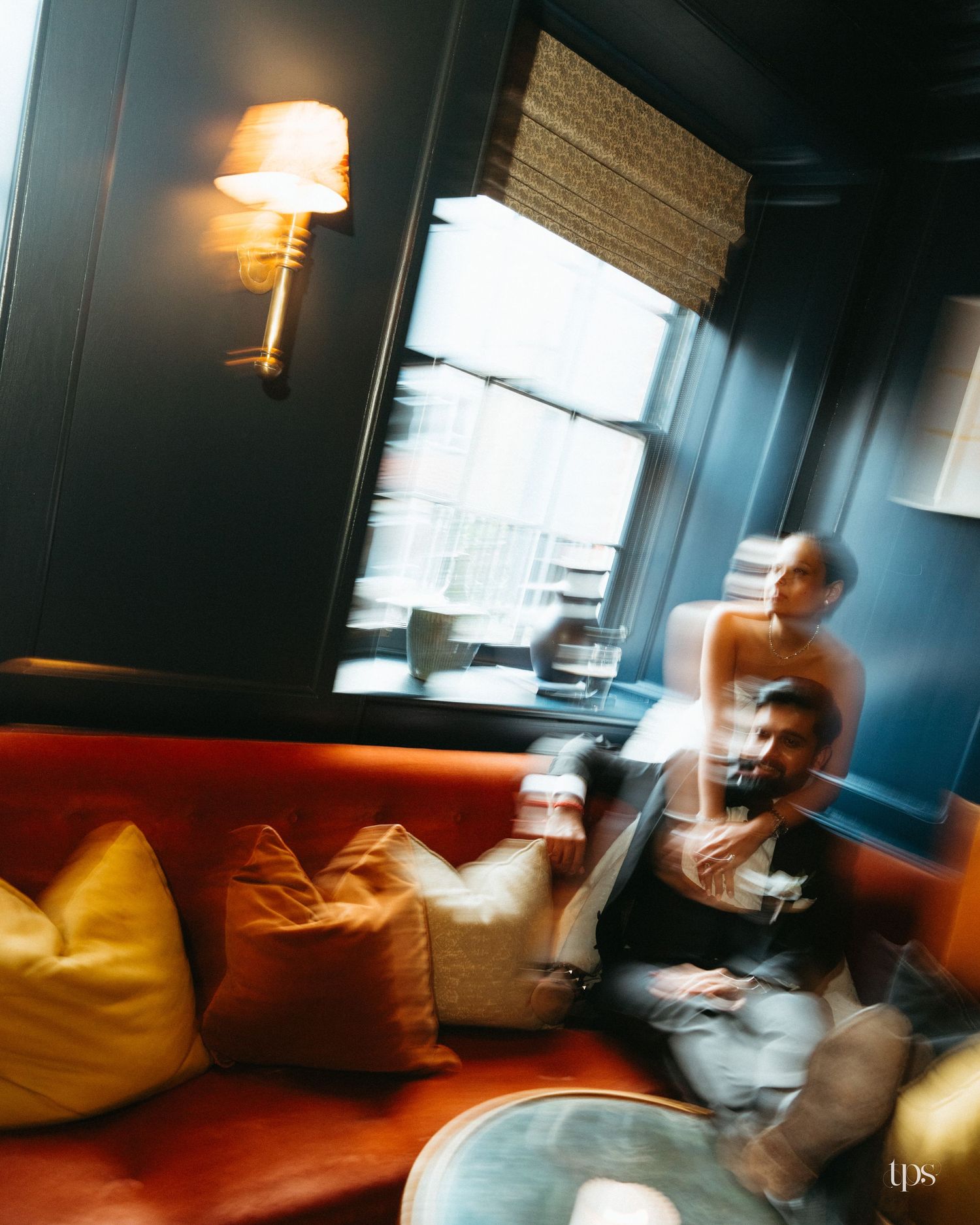 Blurred motion photo of a couple sitting on an orange velvet sofa in a moody, dimly lit lounge with warm lighting.
