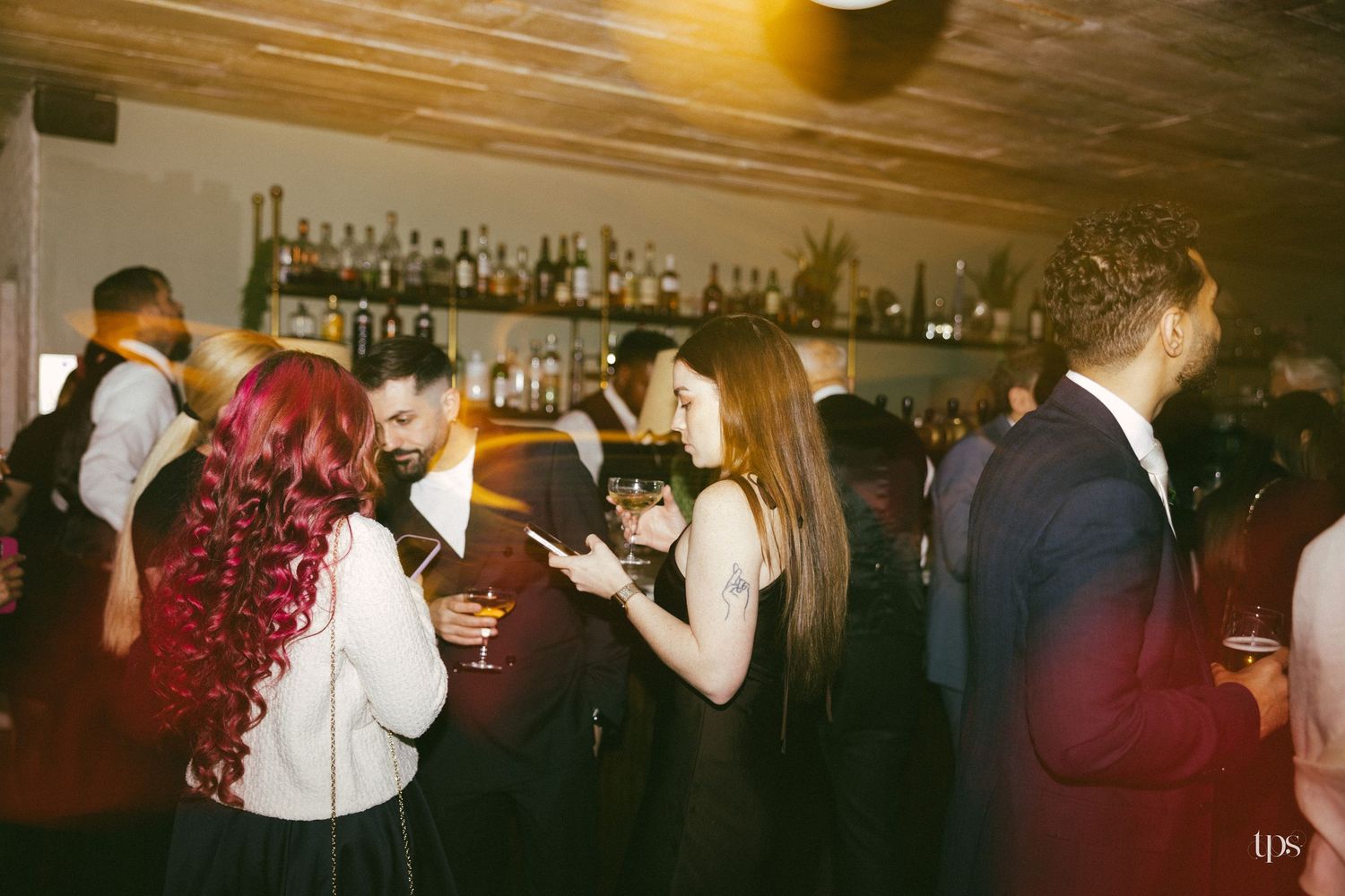 Guests socializing at a dimly lit bar event, with a well-stocked liquor shelf in the background.