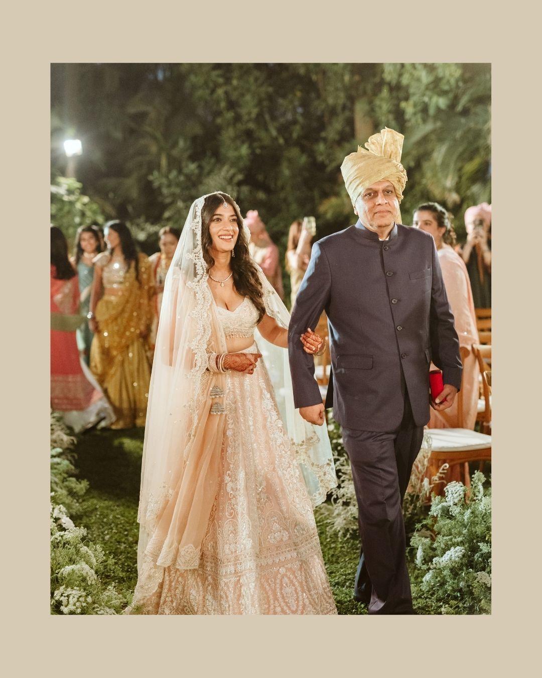 Bride in ivory lehenga walks with elder man in navy sherwani and gold turban at outdoor Indian wedding ceremony.
