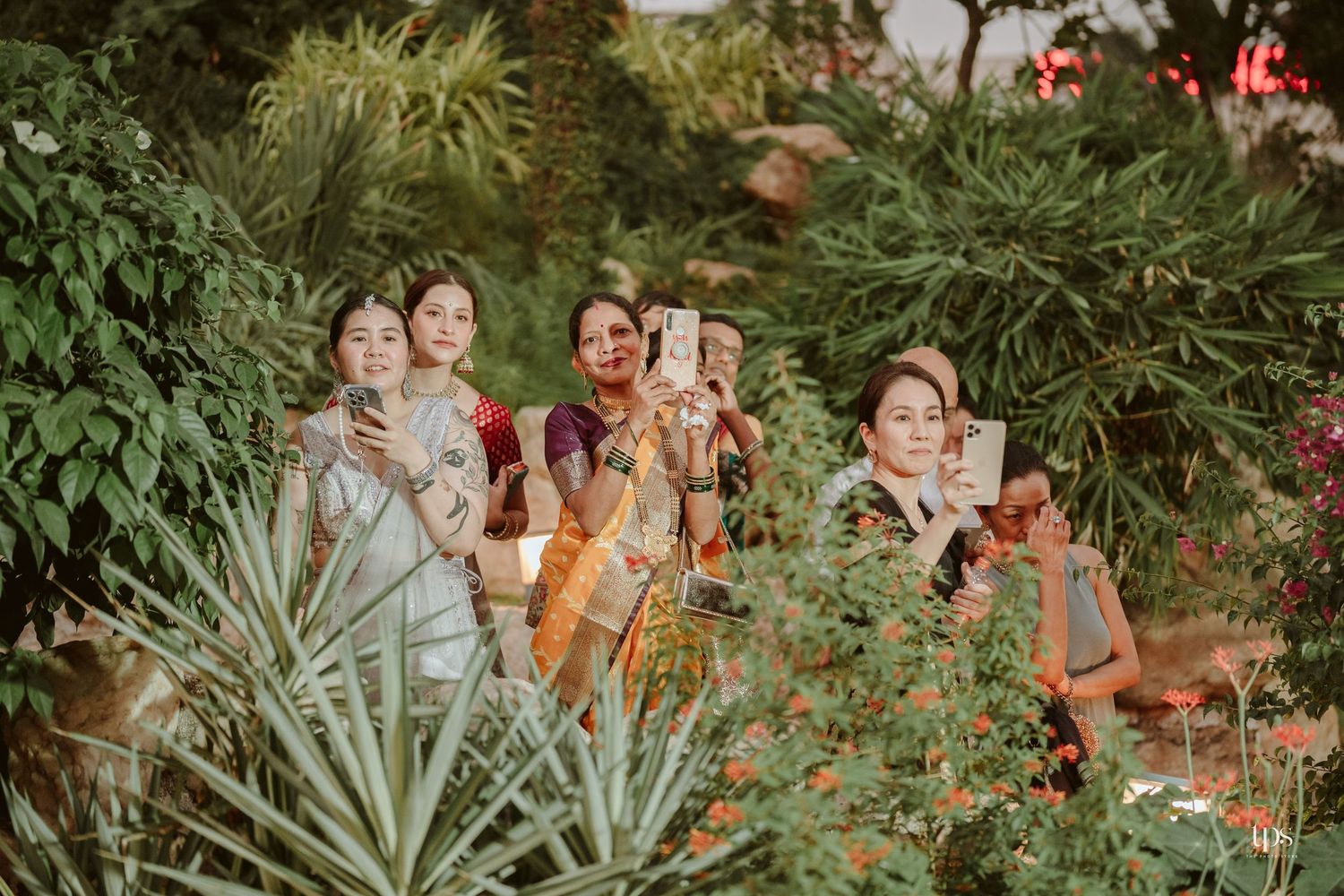 Group of women taking photos with smartphones among lush green plants and red flowers in a garden.