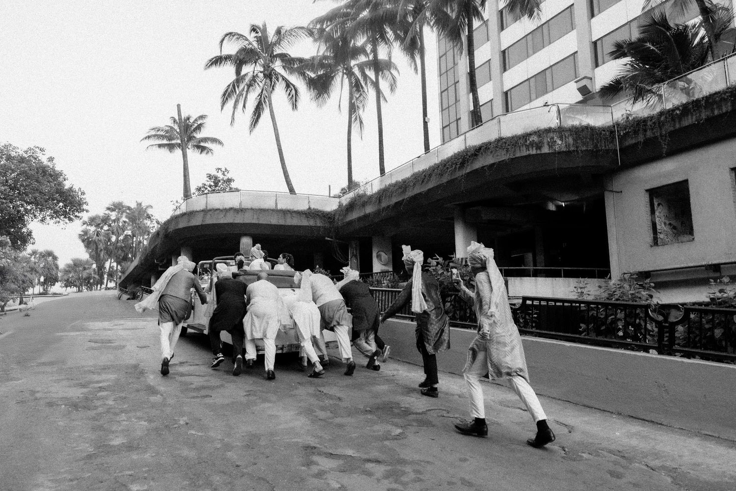 Black and white photo of people pushing a vehicle near a tropical hotel surrounded by palm trees.