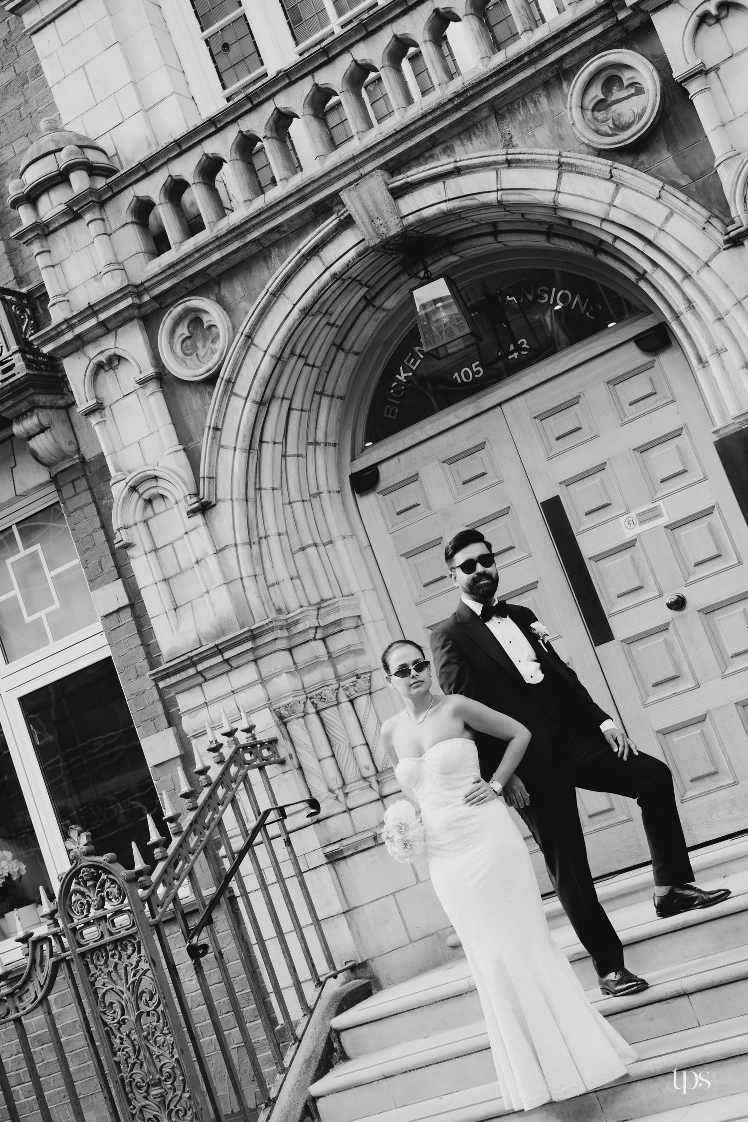 Black and white photo of a bride and groom posing on steps of an elegant stone building in wedding attire.