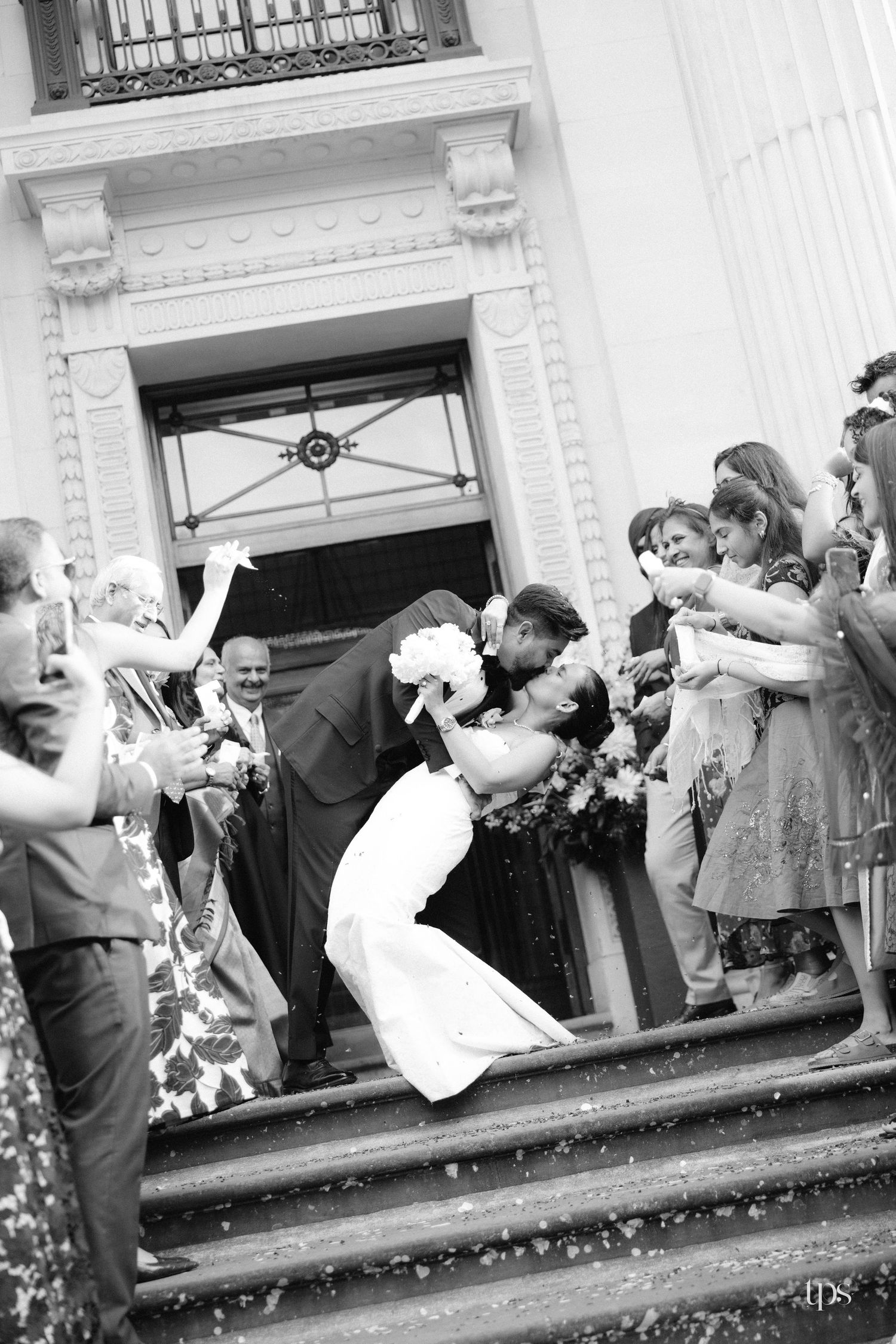 Black and white photo of bride and groom sharing a romantic dip kiss on church steps surrounded by cheering wedding guests.