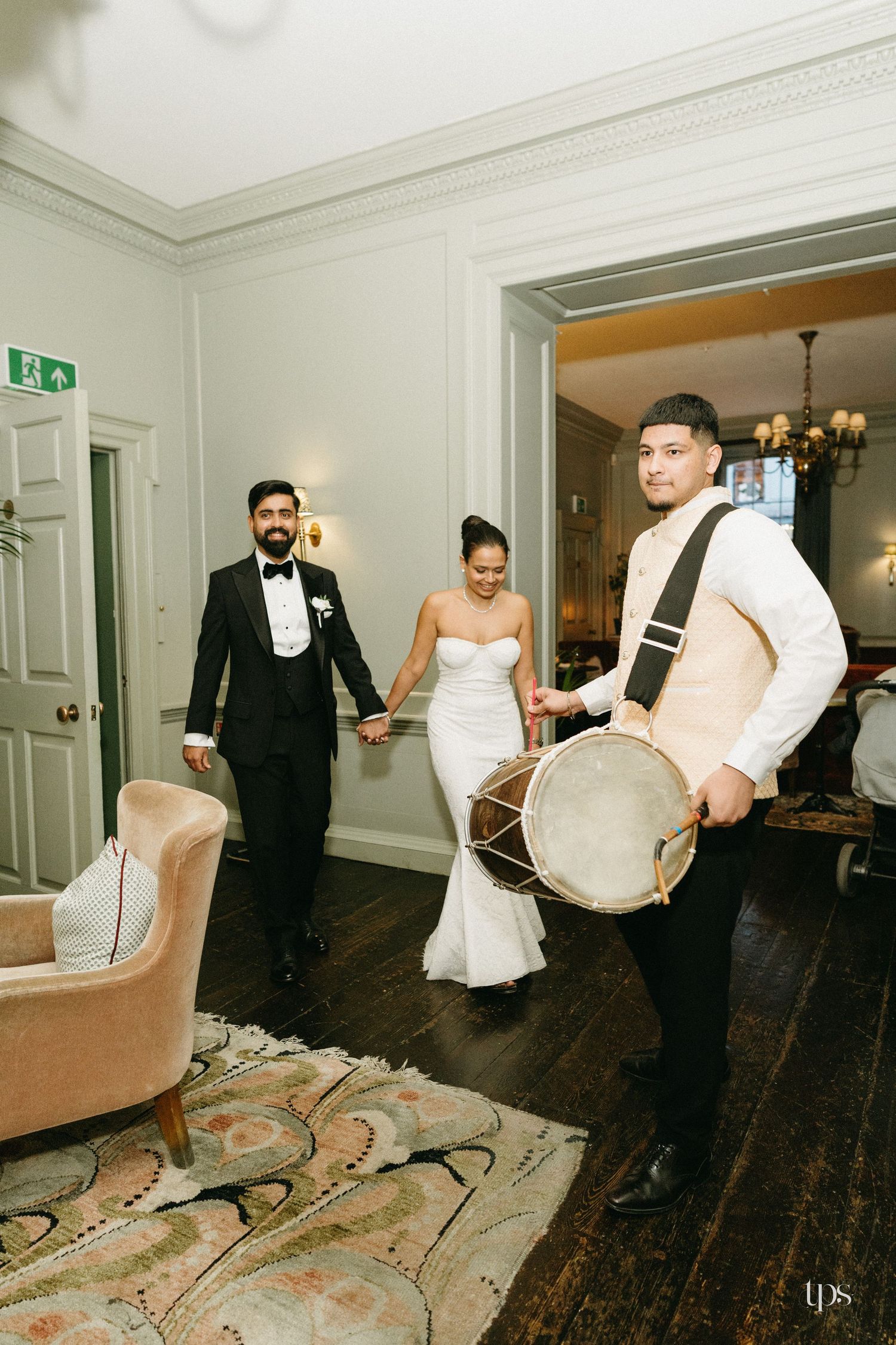 Bride and groom make grand entrance led by dhol drummer at elegant wedding venue with wooden floors.