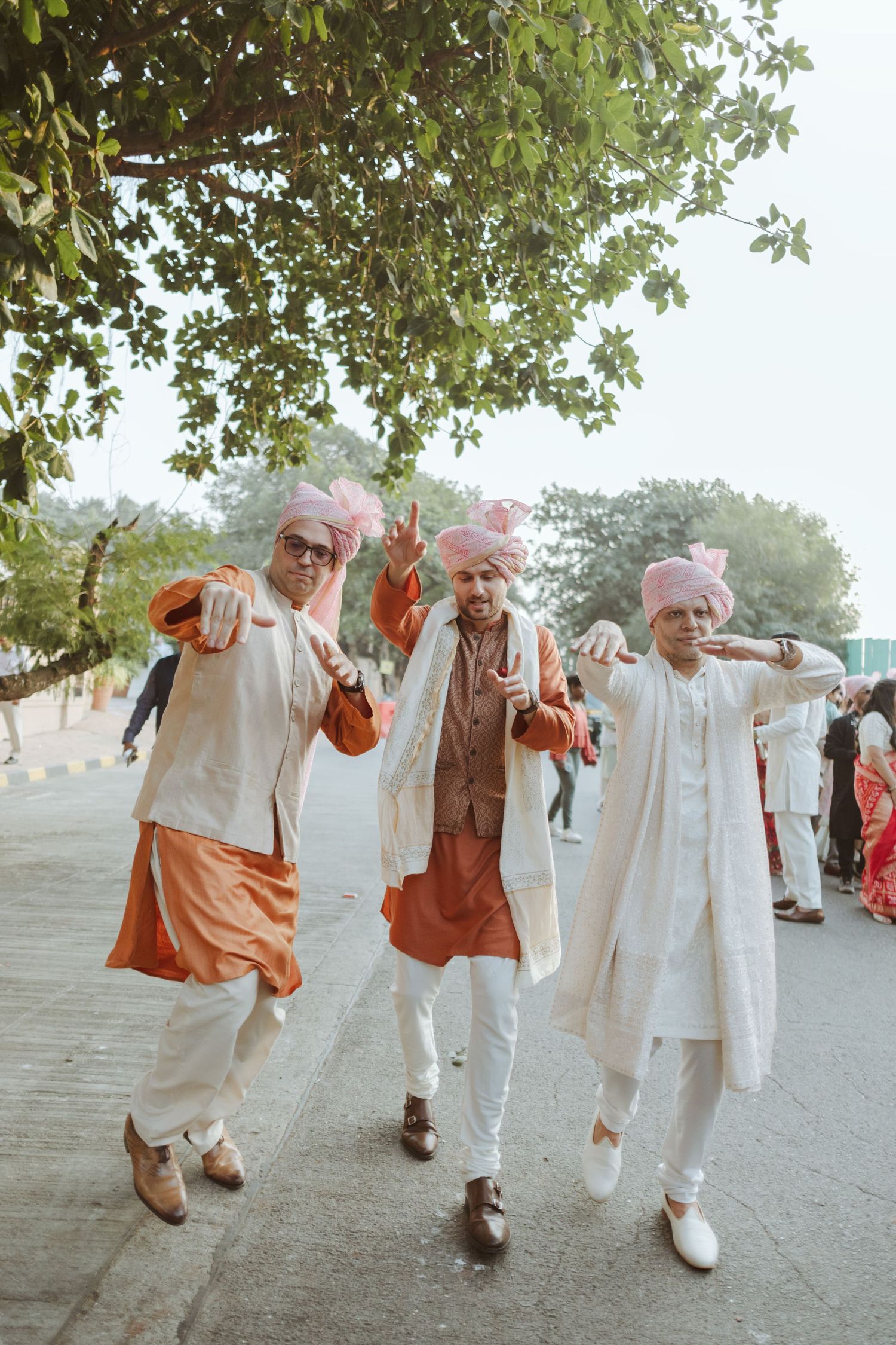 Three men in traditional Indian wedding attire with pink turbans posing outdoors on a sunny day.