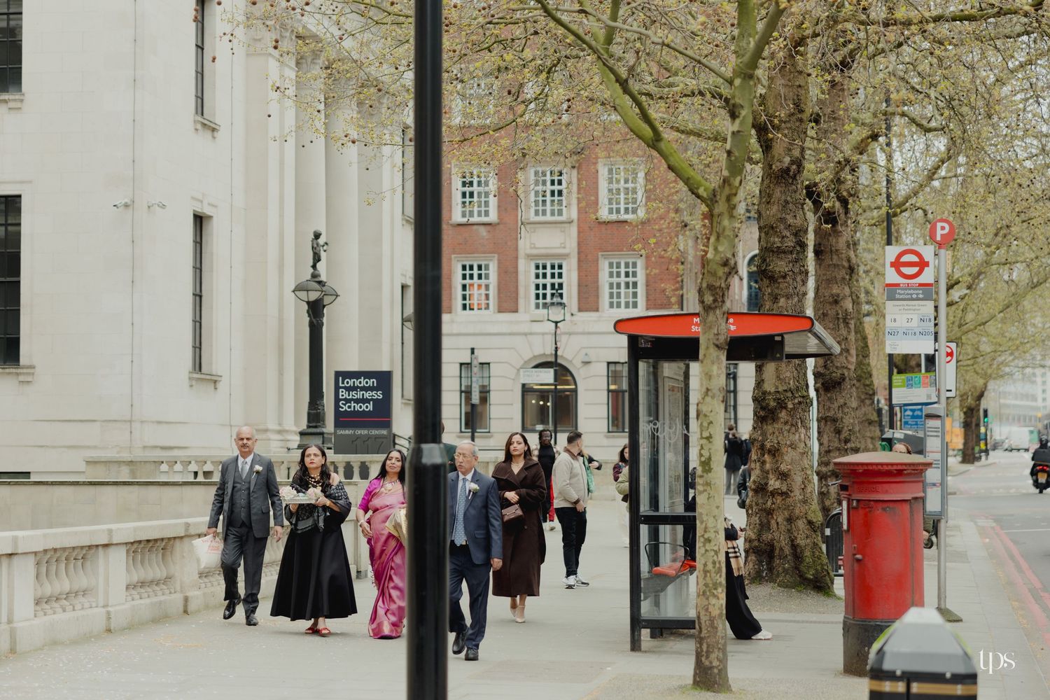 Wedding guests in formal attire walk along a London street near a red phone box and pillar box.