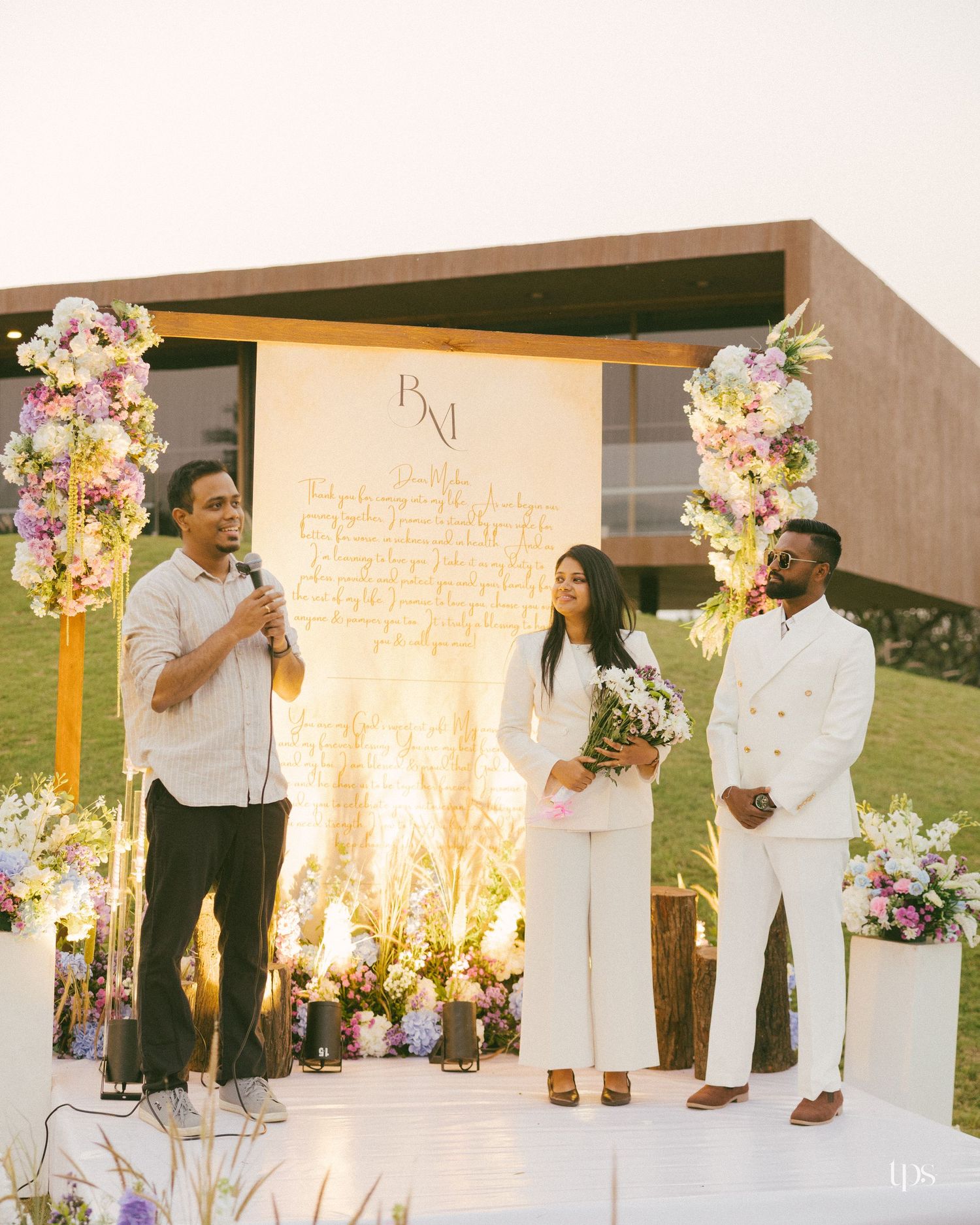 A man speaks into a microphone beside a bride and groom in white outfits at an outdoor wedding ceremony with floral arrangements.