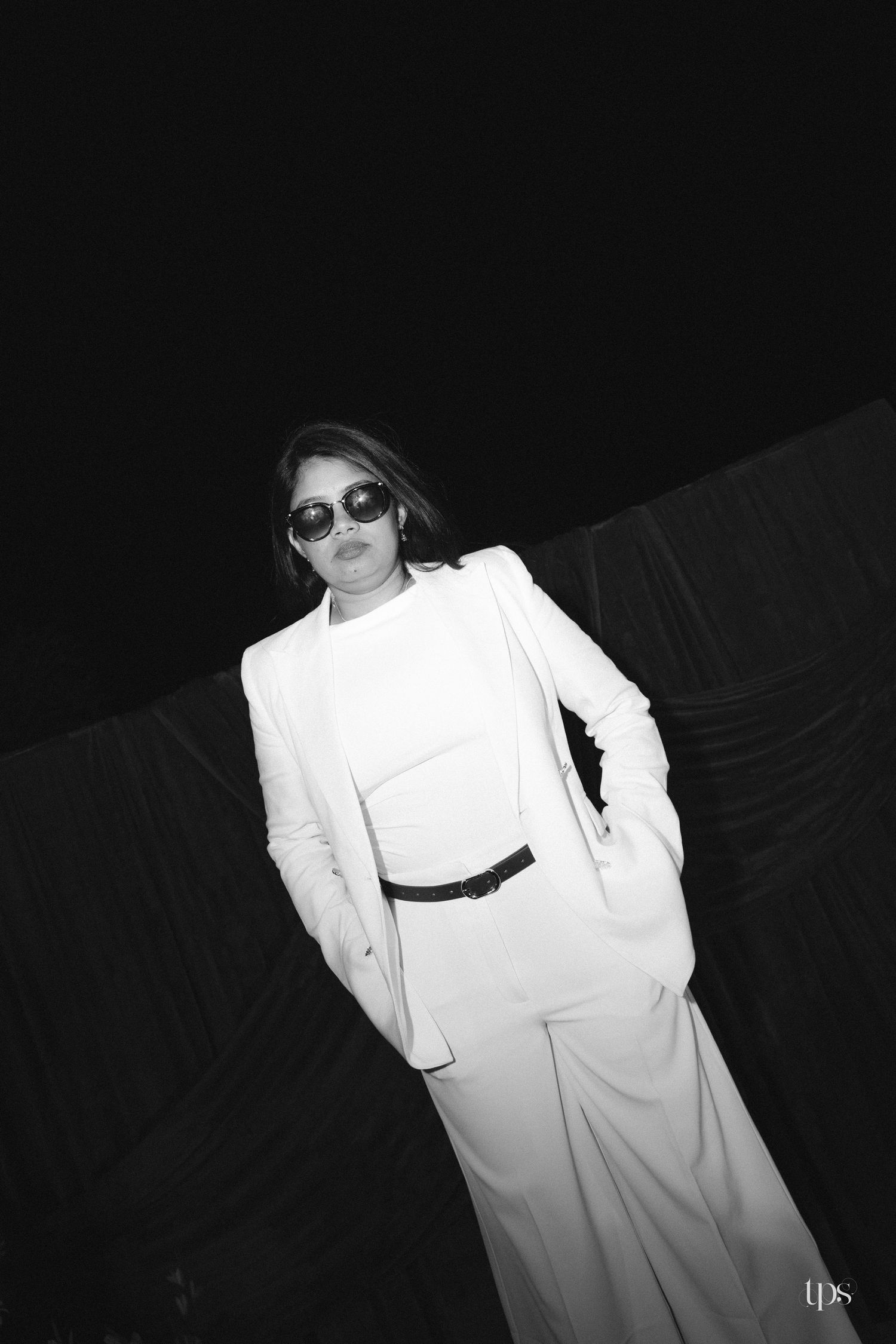 Black and white fashion portrait of a woman in sunglasses, white blazer, top and wide-leg trousers with a black belt against a dark backdrop