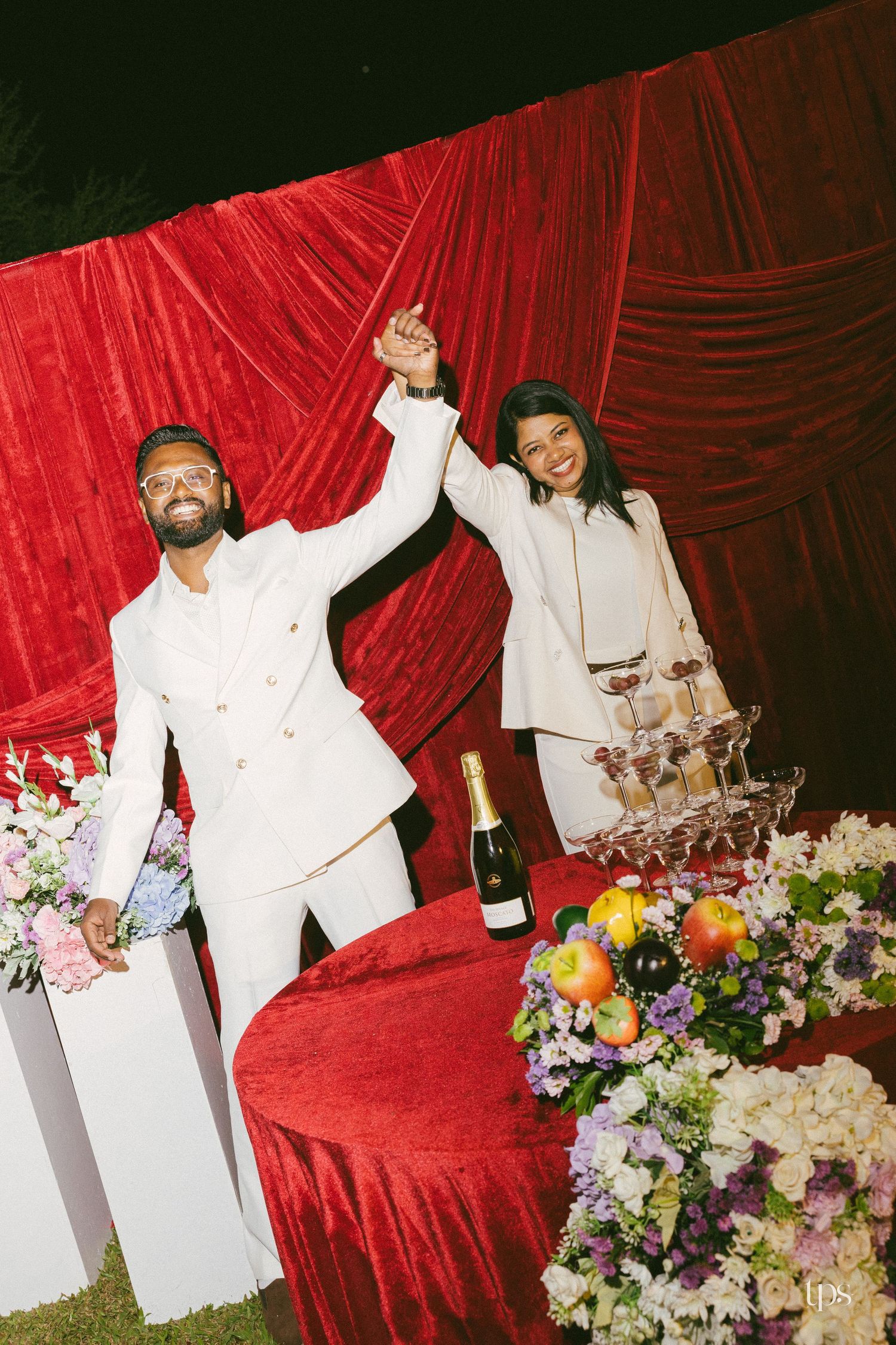 Couple in white suits raise joined hands beside a champagne tower and fruit and floral centerpiece against a red velvet backdrop.