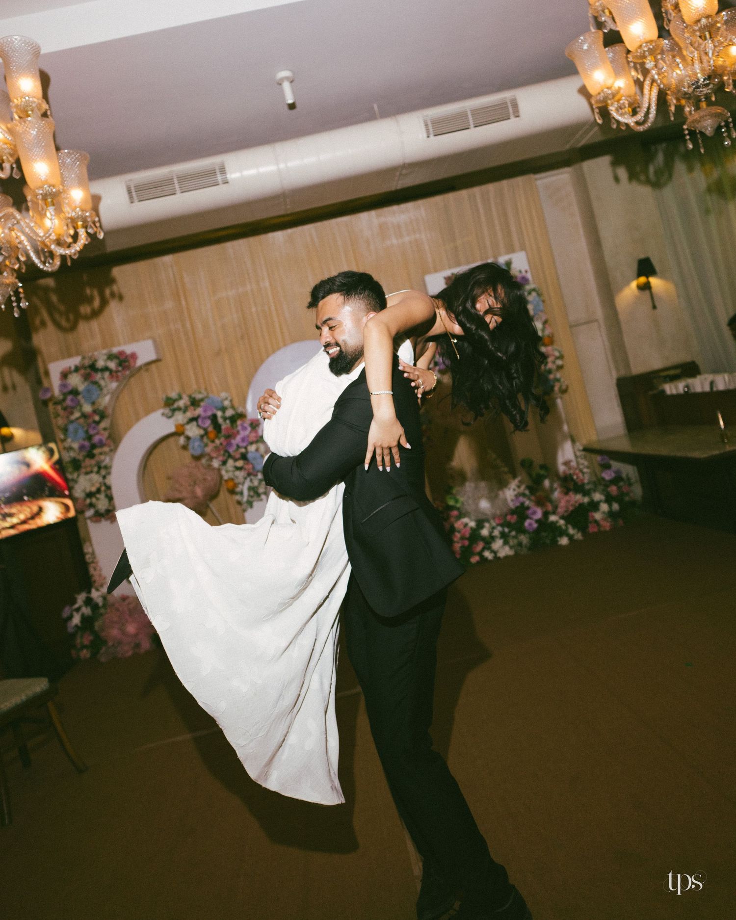 Groom in a black suit lifts a bride in a flowing white gown during a wedding dance beneath chandeliers and floral decor.