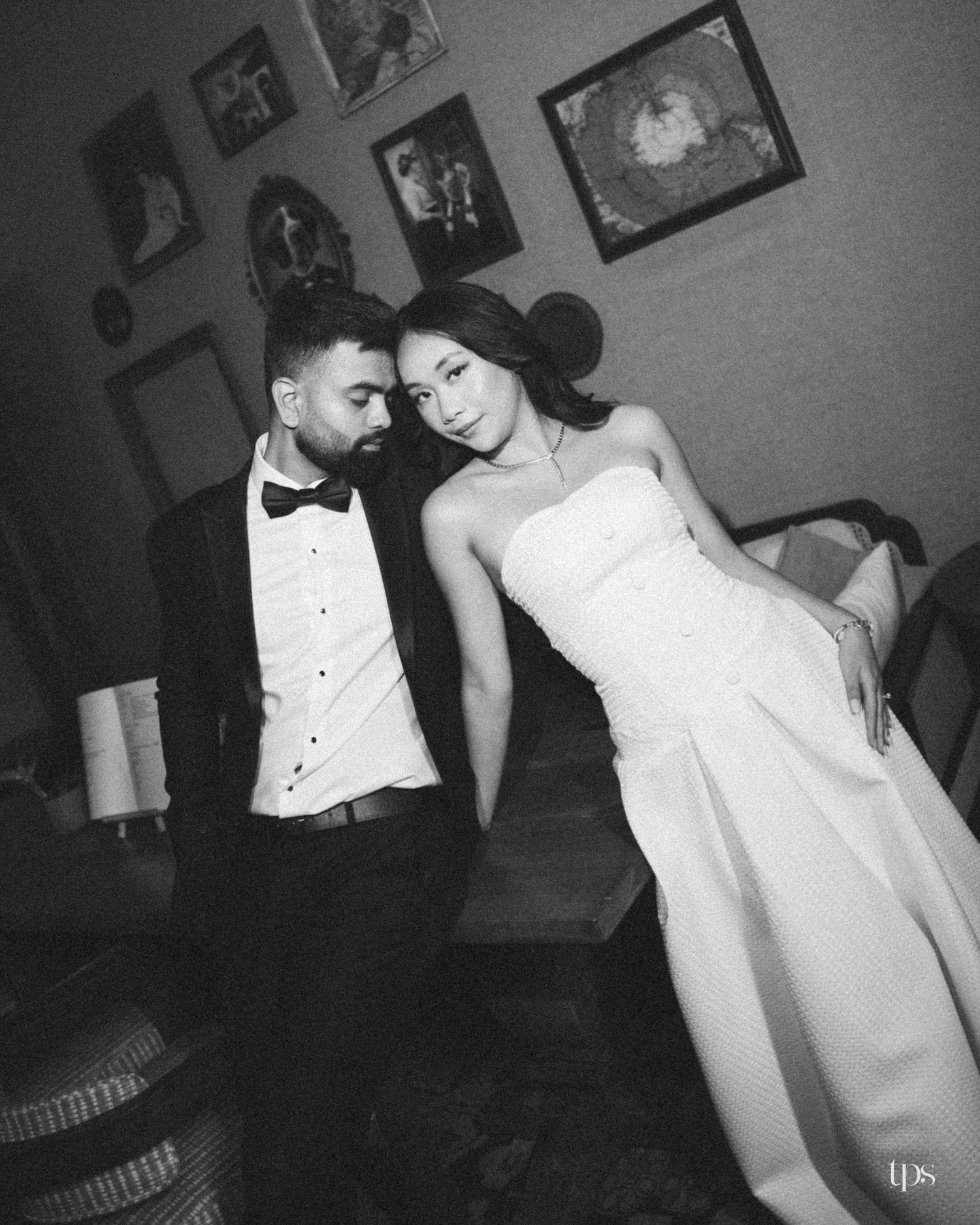 Black and white portrait of a couple in formal attire, a man in a tuxedo and bow tie beside a woman in a strapless gown, framed art behind.