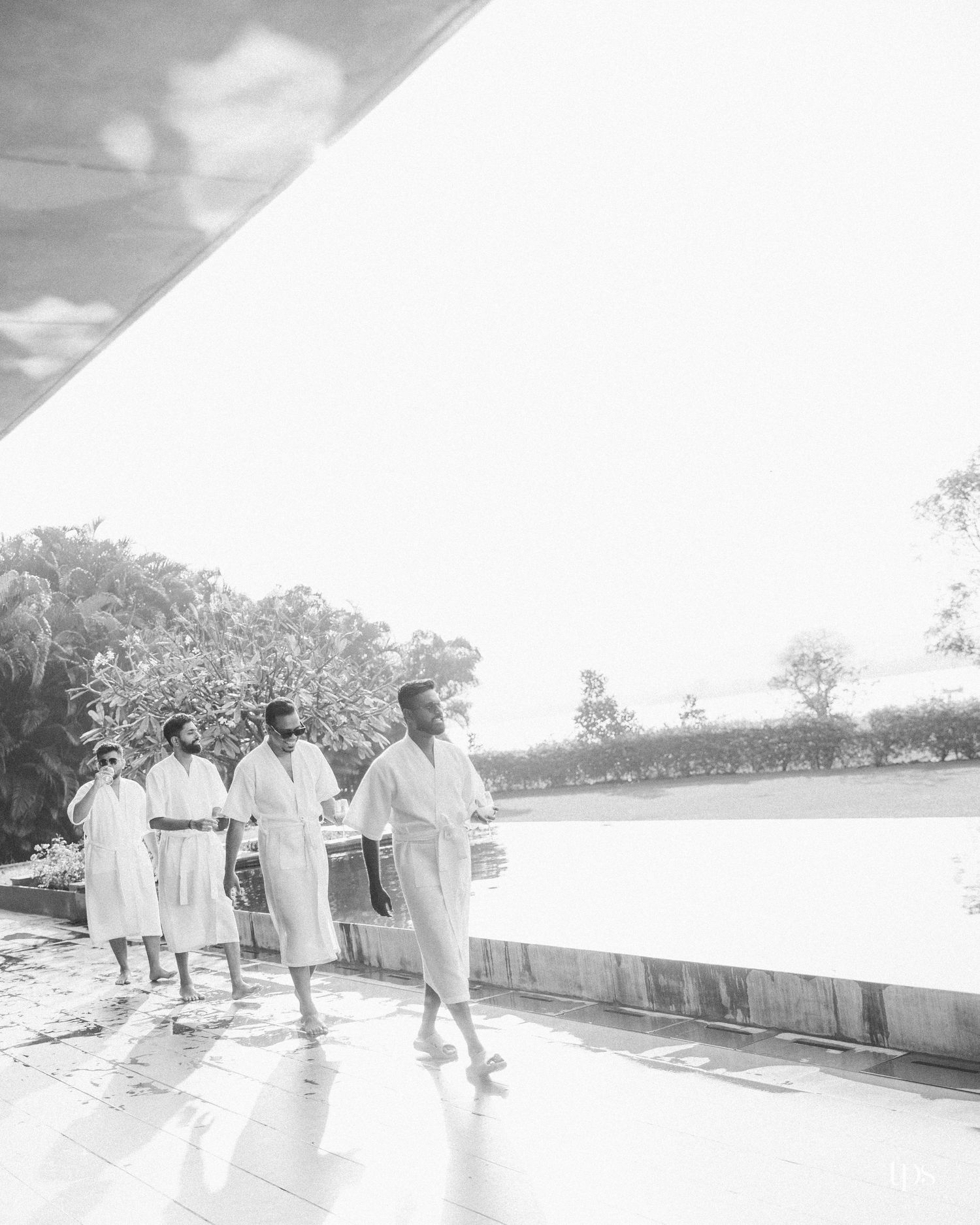 Four men in white robes walk single file beside a sunlit infinity pool with lush tropical trees behind them in a black and white photo.