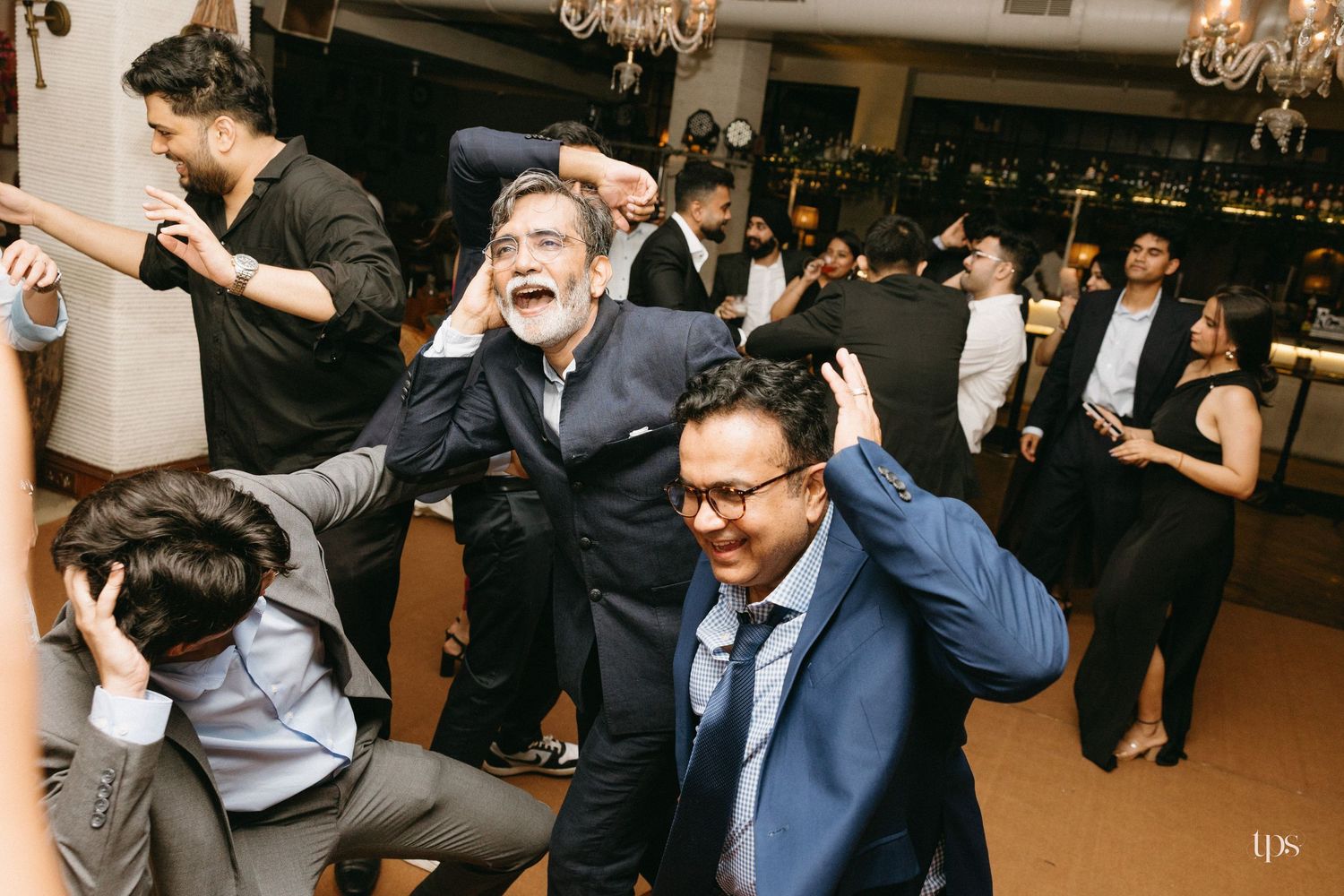 Group of men in suits dancing and laughing together at a lively indoor party with chandeliers and guests in the background.