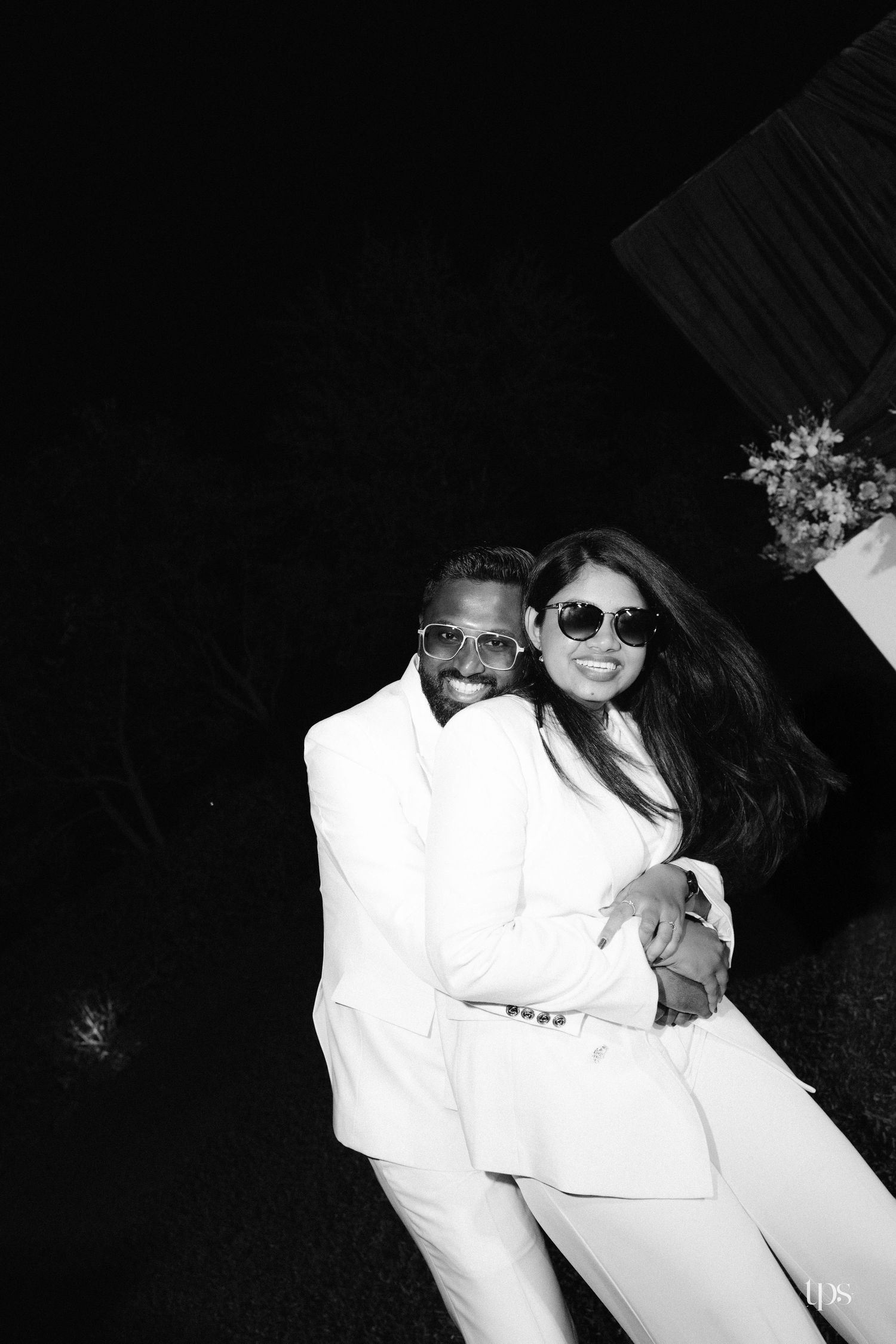 Black and white photo of a smiling couple in white suits and sunglasses embracing outdoors at night, with flowers visible in the corner.