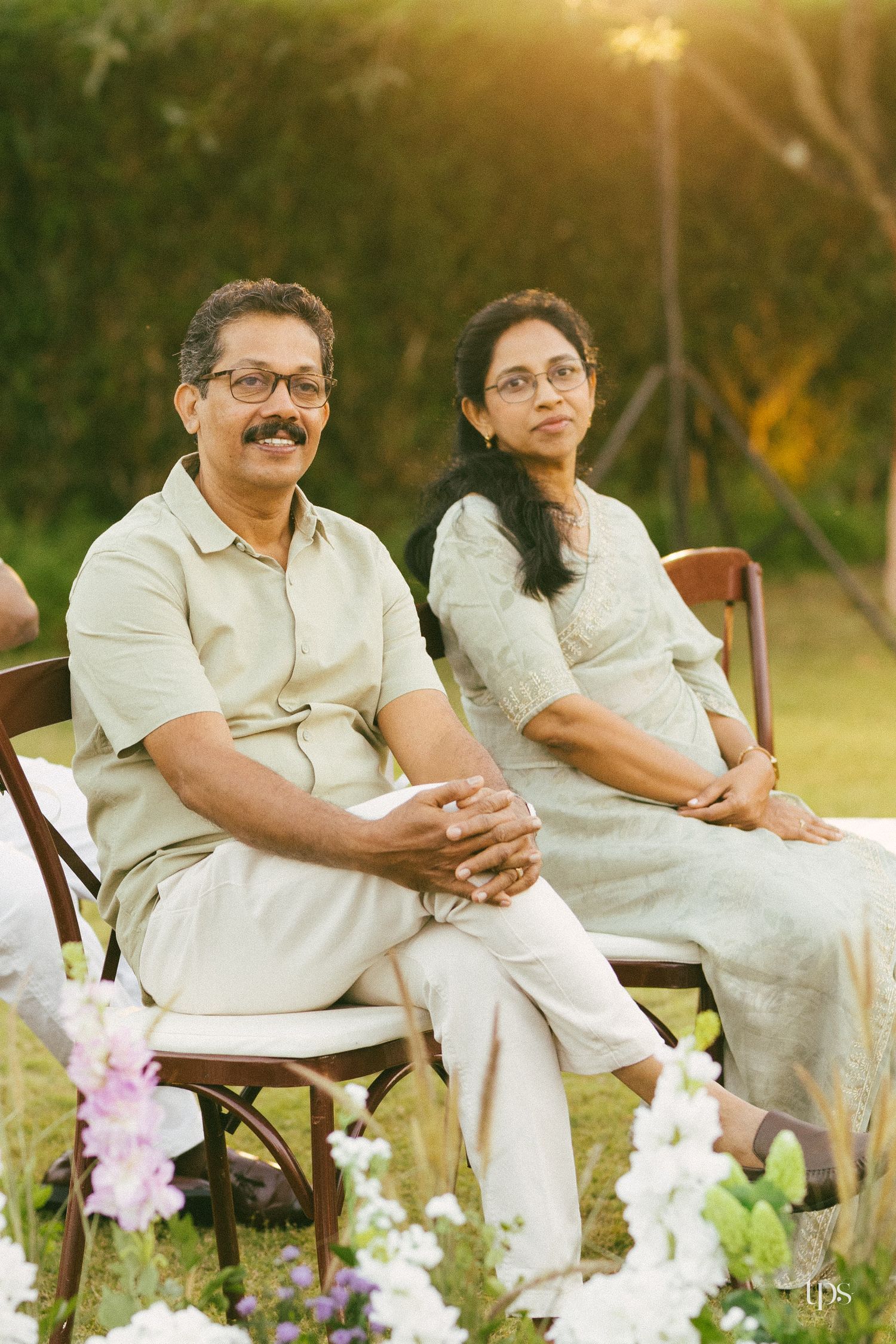 A smiling man and woman in light, coordinated outfits sit on wooden chairs at an outdoor garden ceremony with flowers in the foreground.