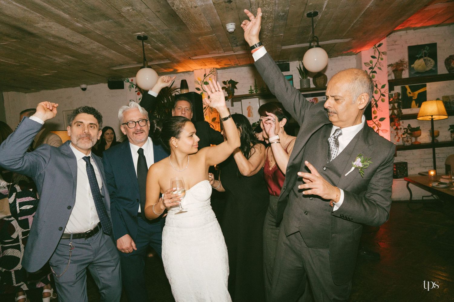 Bride in white dress celebrates with guests on wedding reception dance floor, arms raised in joy.