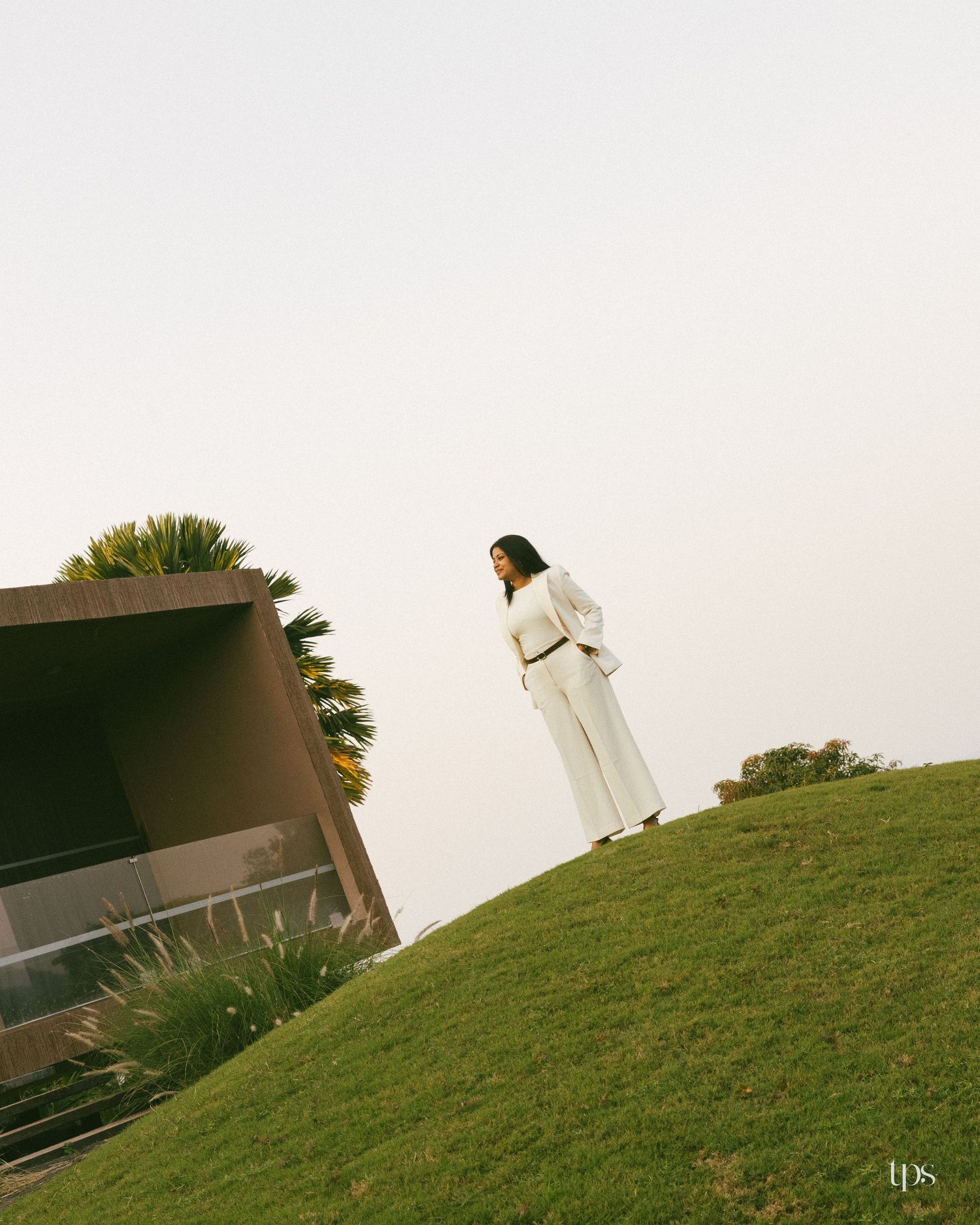 Woman in a white blazer and wide-leg pants stands on a grassy hill beside a modern concrete building and palm tree under a hazy sky.
