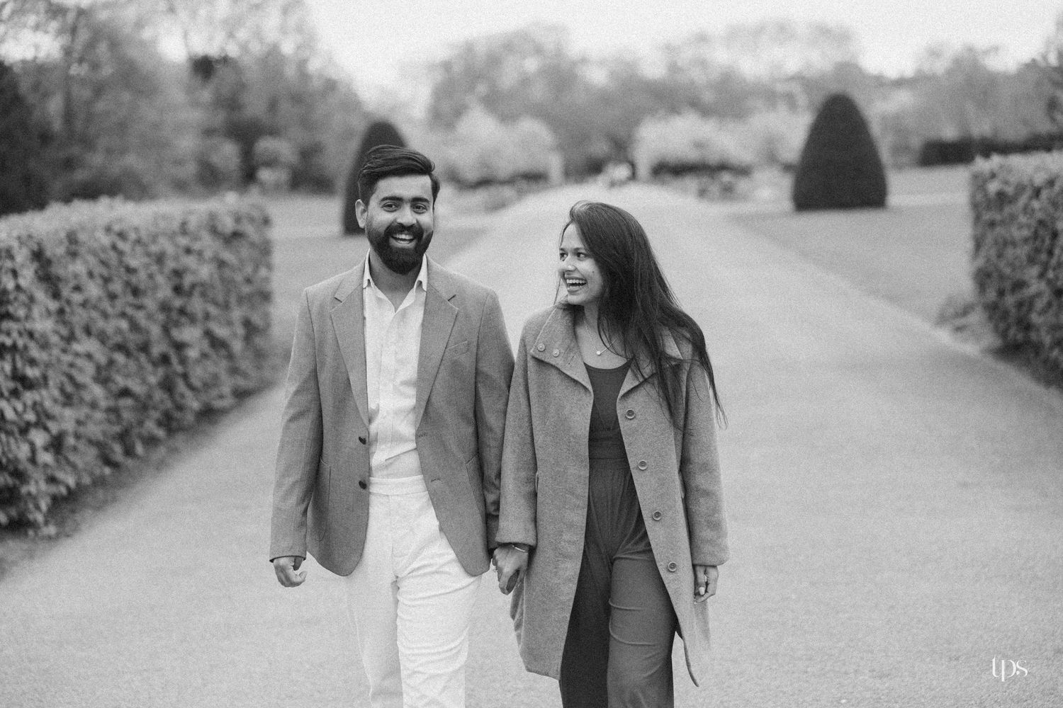 Couple walking hand in hand along a tree-lined garden path in a black and white engagement photo.