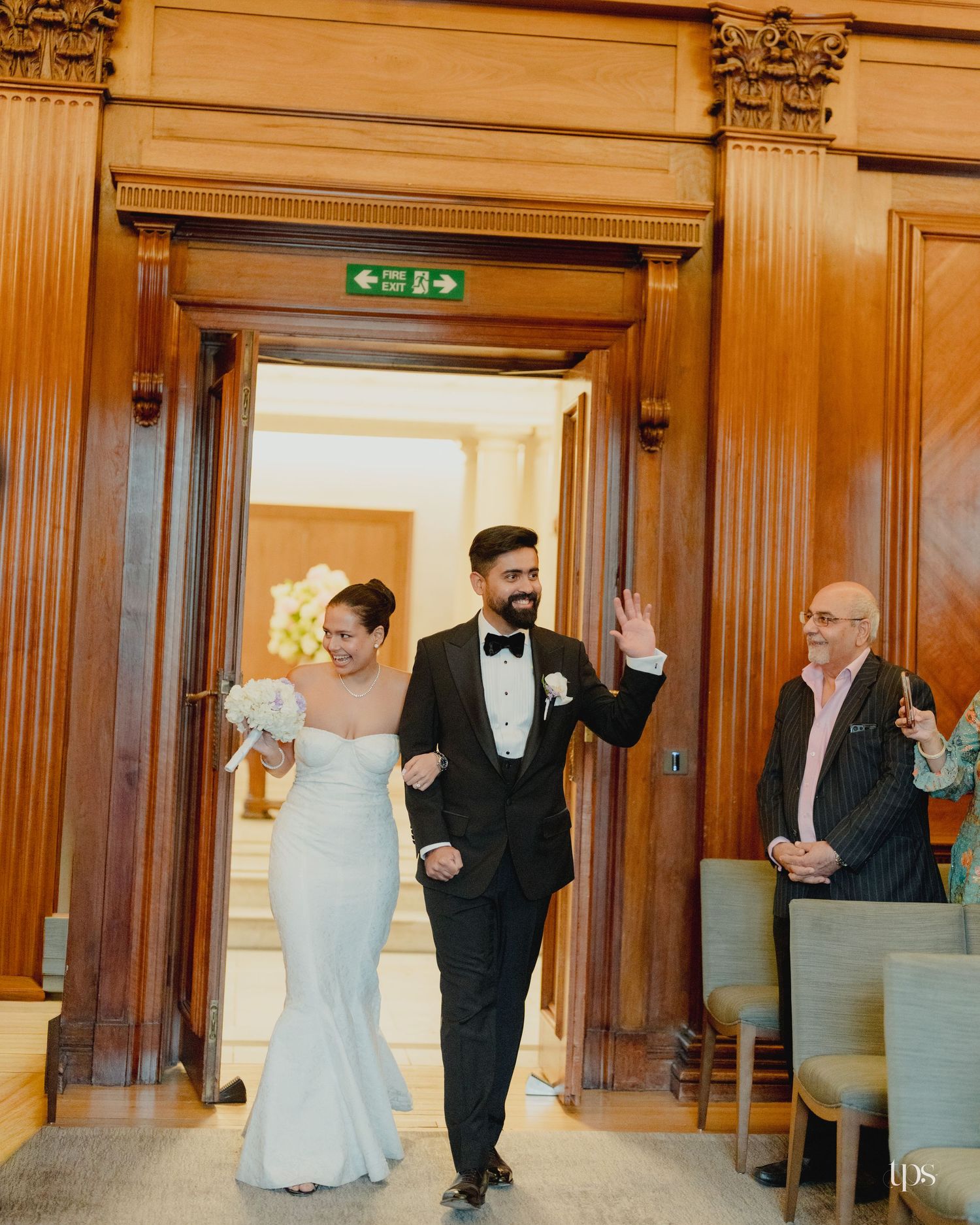 Bride and groom walk arm-in-arm through ornate wooden doorway, she in white gown, he in black tuxedo, at elegant venue.