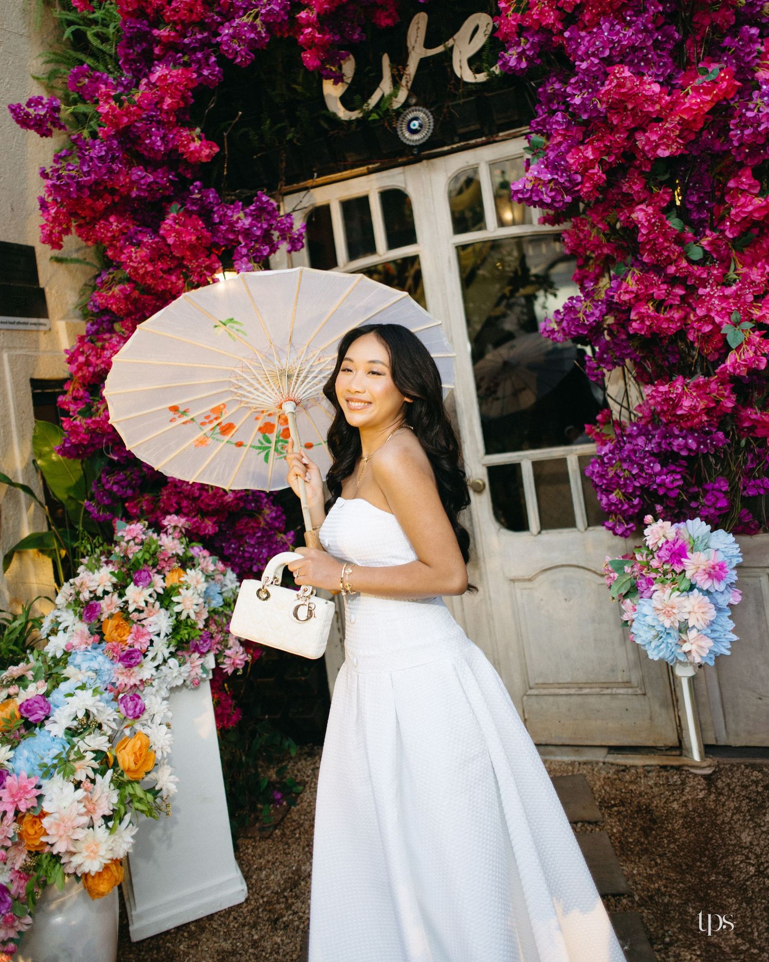 Woman in a white strapless gown holding a floral paper parasol and white handbag, standing before a door framed by pink bougainvillea.