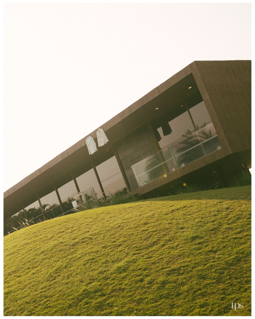 Modern cantilevered building with a concrete roof and glass walls, perched above a sunlit, manicured green grass hill.