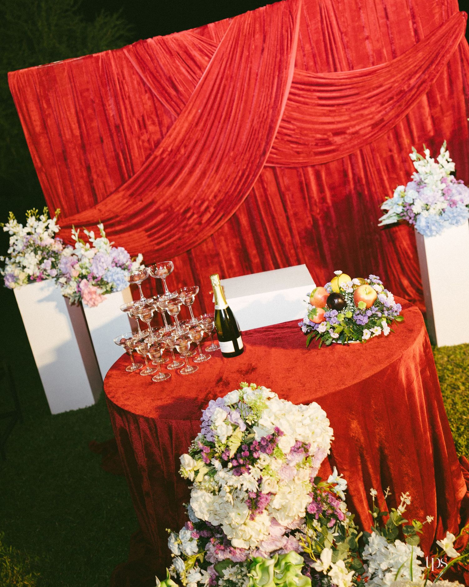 Red velvet draped table with a champagne glass tower and bottle, surrounded by white and lavender floral arrangements at an outdoor wedding.