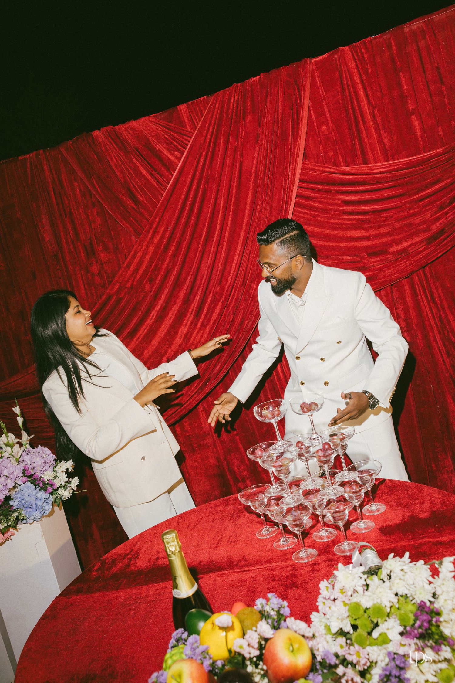 Two people in white suits laugh beside a champagne tower on a red velvet-draped table with fruit and flowers.
