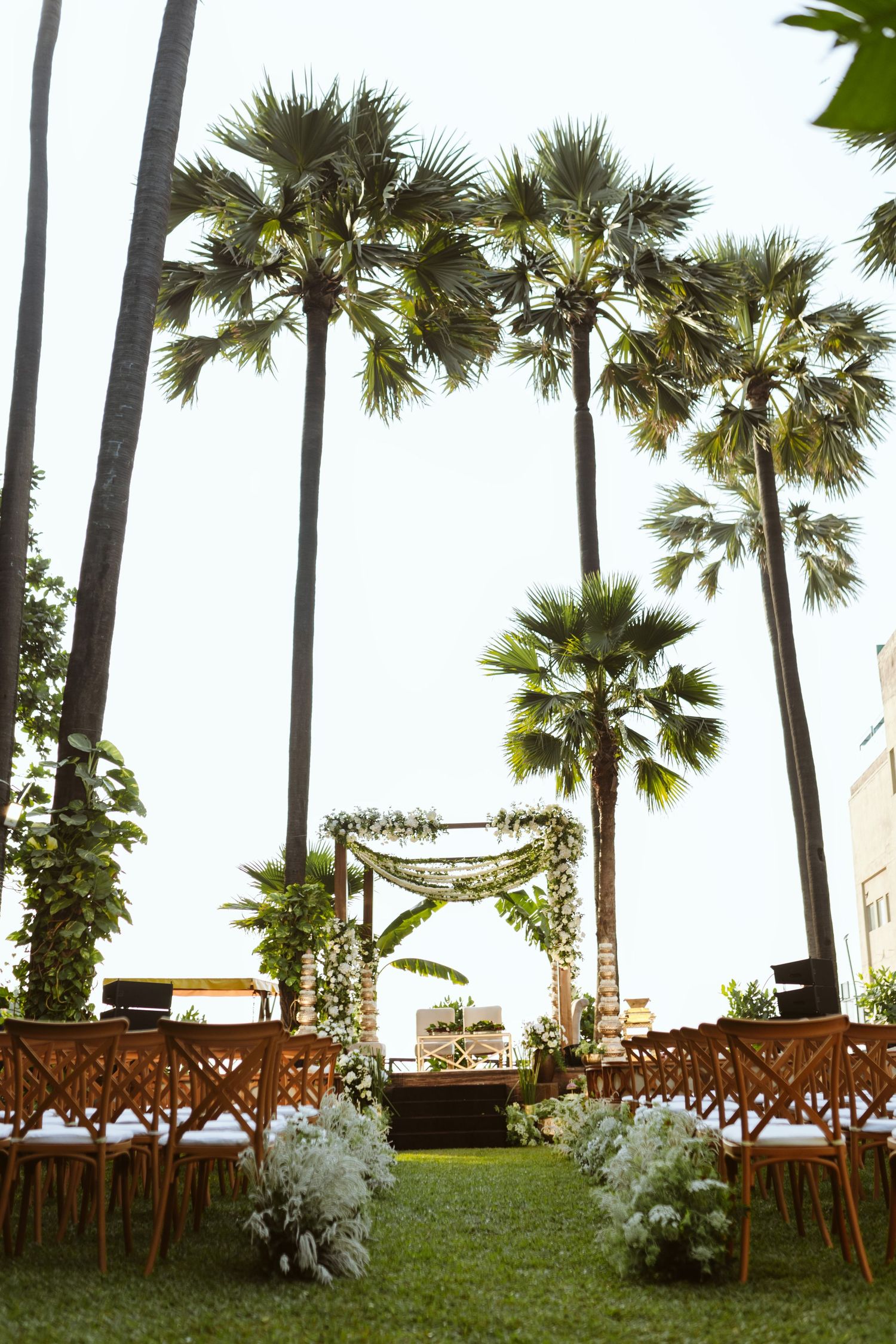Outdoor wedding ceremony setup with wooden chairs, floral arch, and tall palm trees on green lawn.