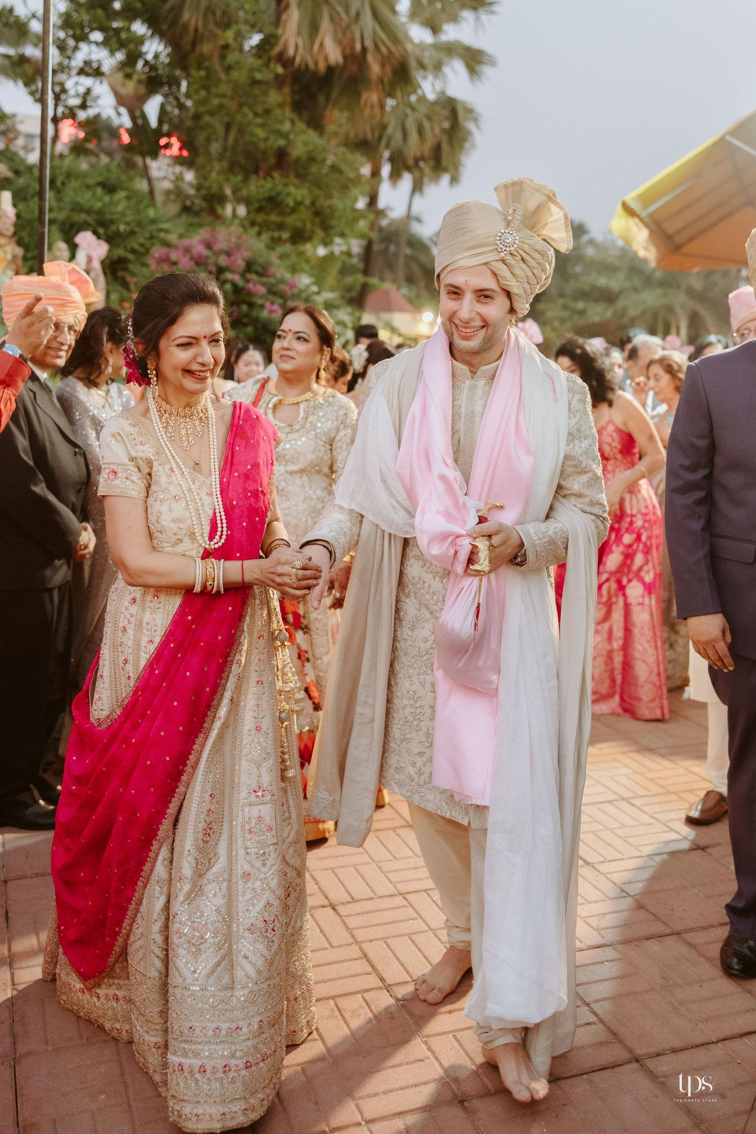 Bride in pink and gold lehenga walks with groom in cream sherwani and turban at traditional Indian wedding ceremony.