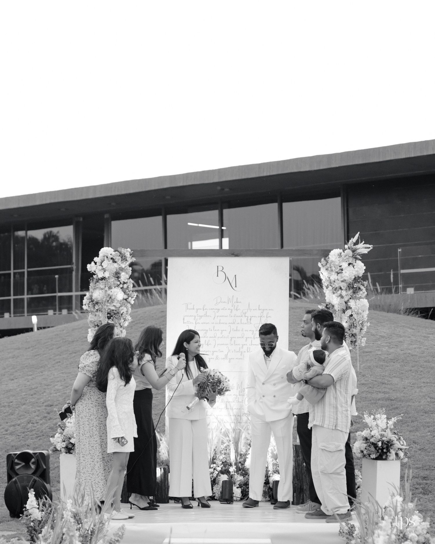 Black and white photo of a wedding ceremony with a couple and guests gathered before a monogram backdrop and floral arrangements.
