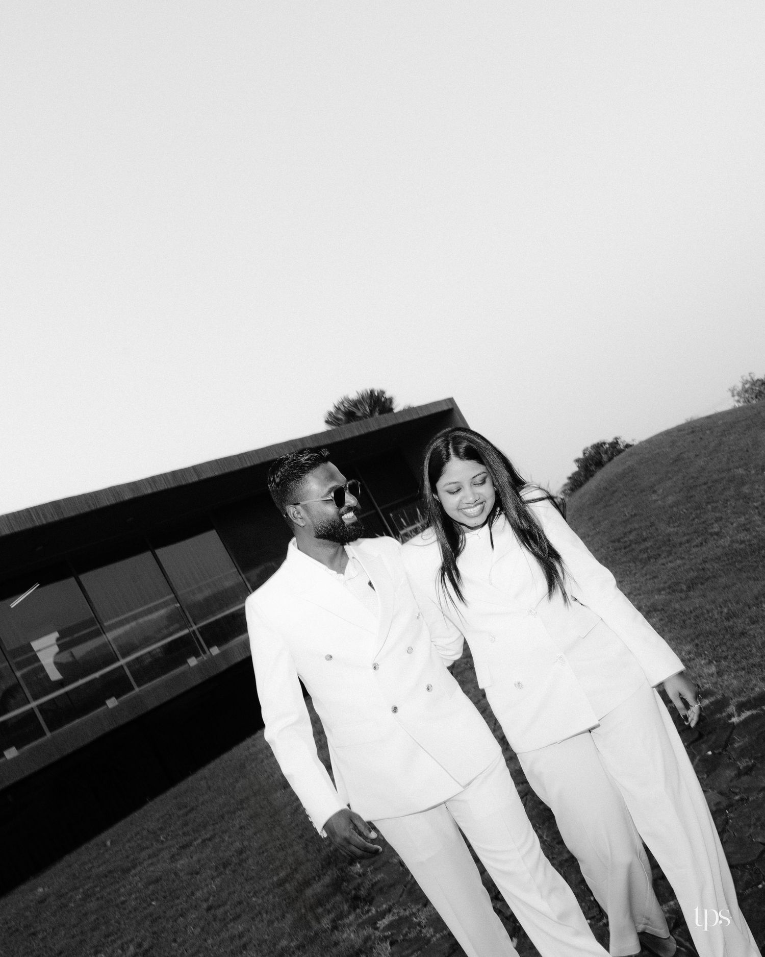 Black and white photo of a smiling couple in matching white suits walking arm in arm on a grassy hill near a modern building.