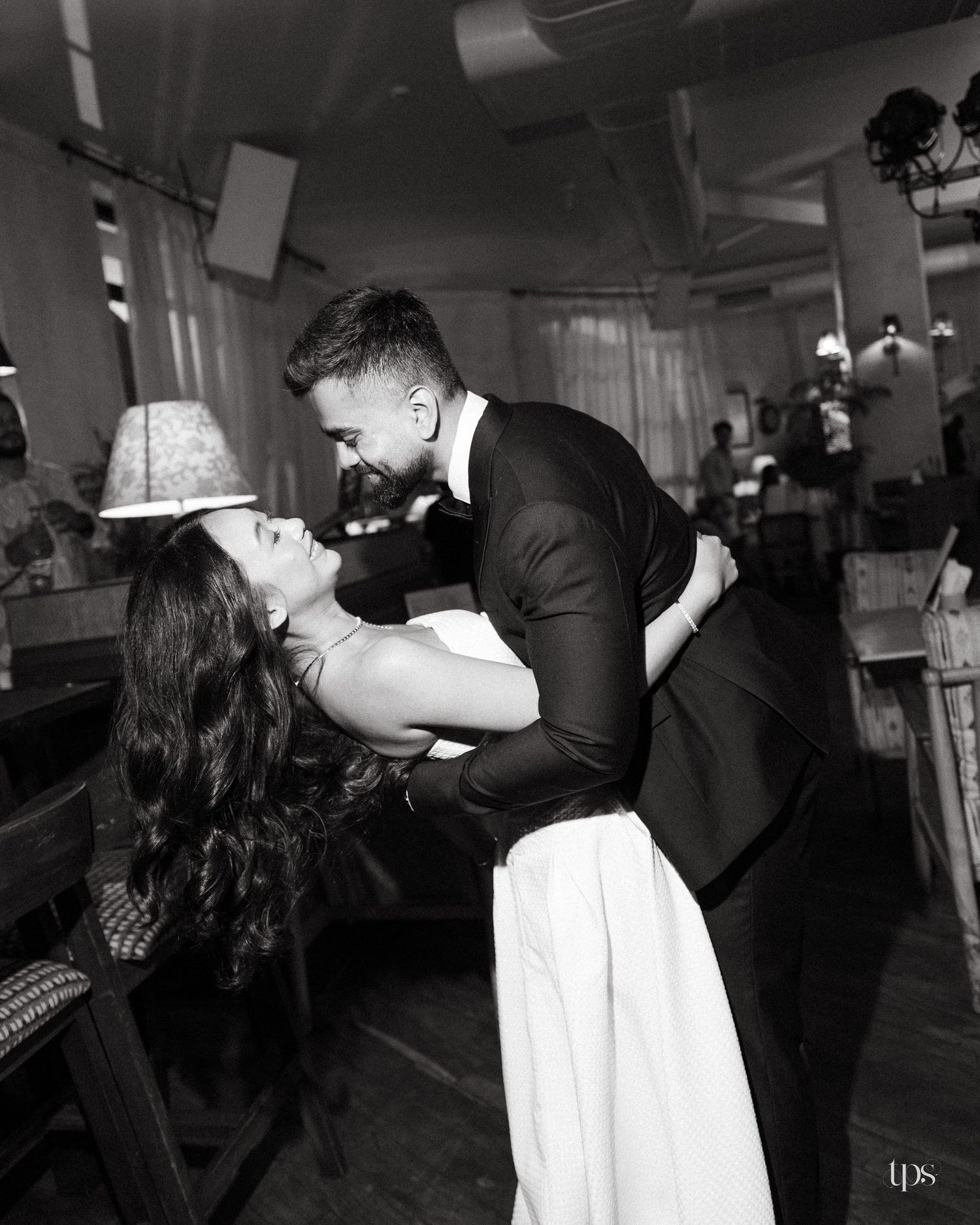 Black and white photo of a groom in a dark suit dipping a bride in a white dress during a romantic dance in a cozy venue.