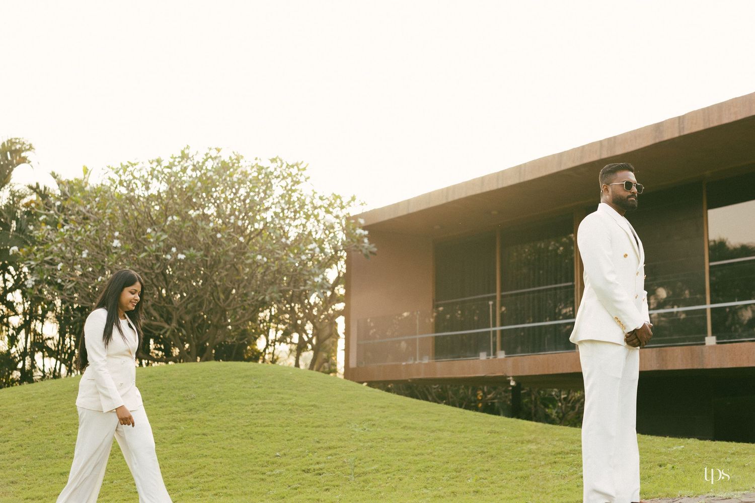 Man and woman in matching white outfits stand apart on a grassy hill in front of a modern building with large glass windows.