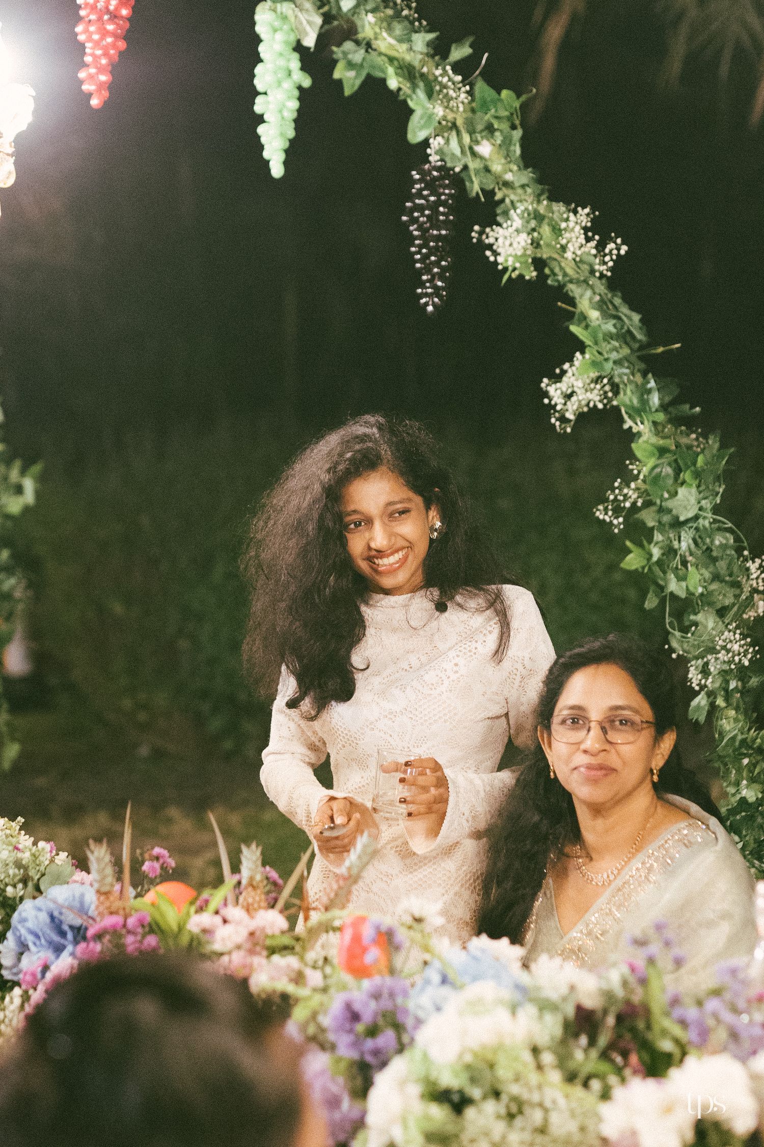 Two smiling women in elegant cream outfits stand behind colorful flower arrangements under a leafy floral arch at a night event.